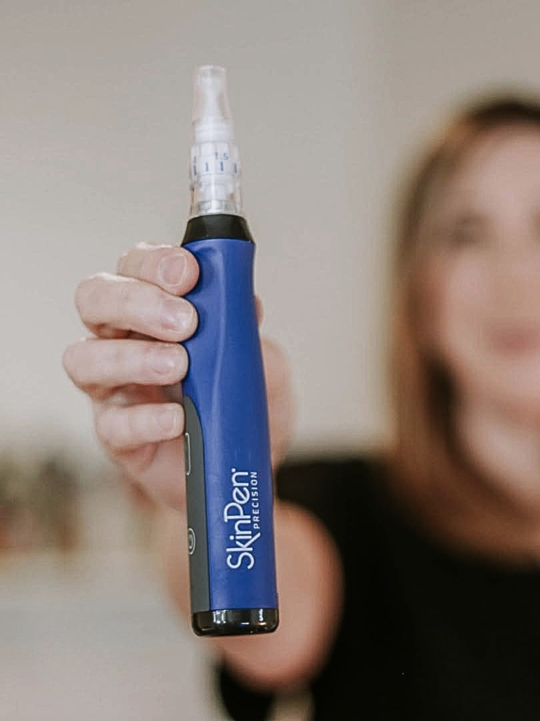 A person holding a blue SkinPen precision device with a clear tip, with a blurred background and a woman smiling to the right.