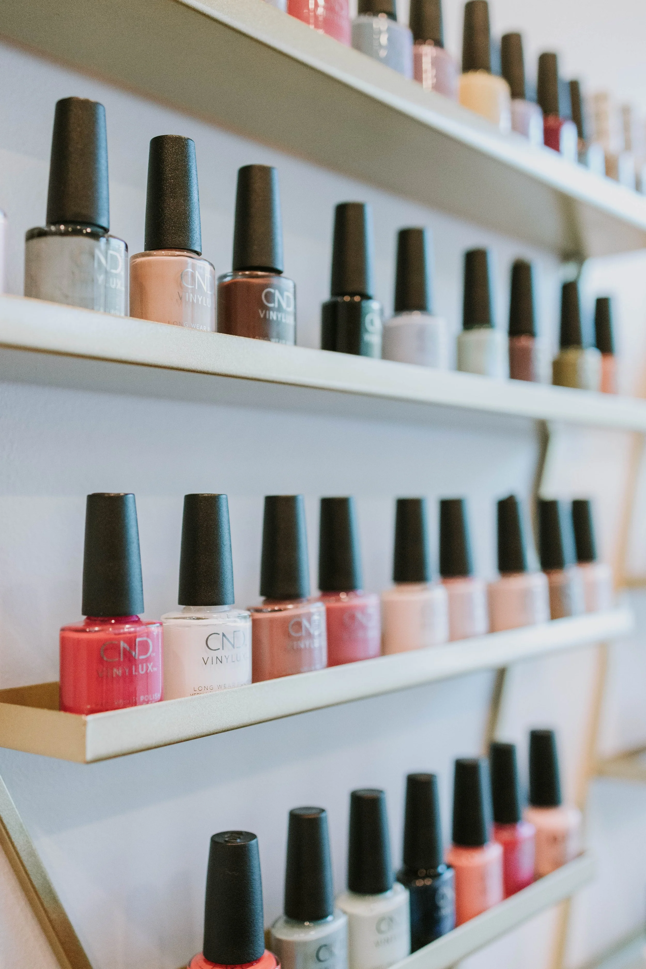 A display shelf with rows of nail polish bottles in various colors including pink, white, beige, brown, and black, with some bottles featuring black caps and others with clear caps. The shelf is made of light wood or white material with a angled design, and additional bottles are visible on a higher shelf in the background.