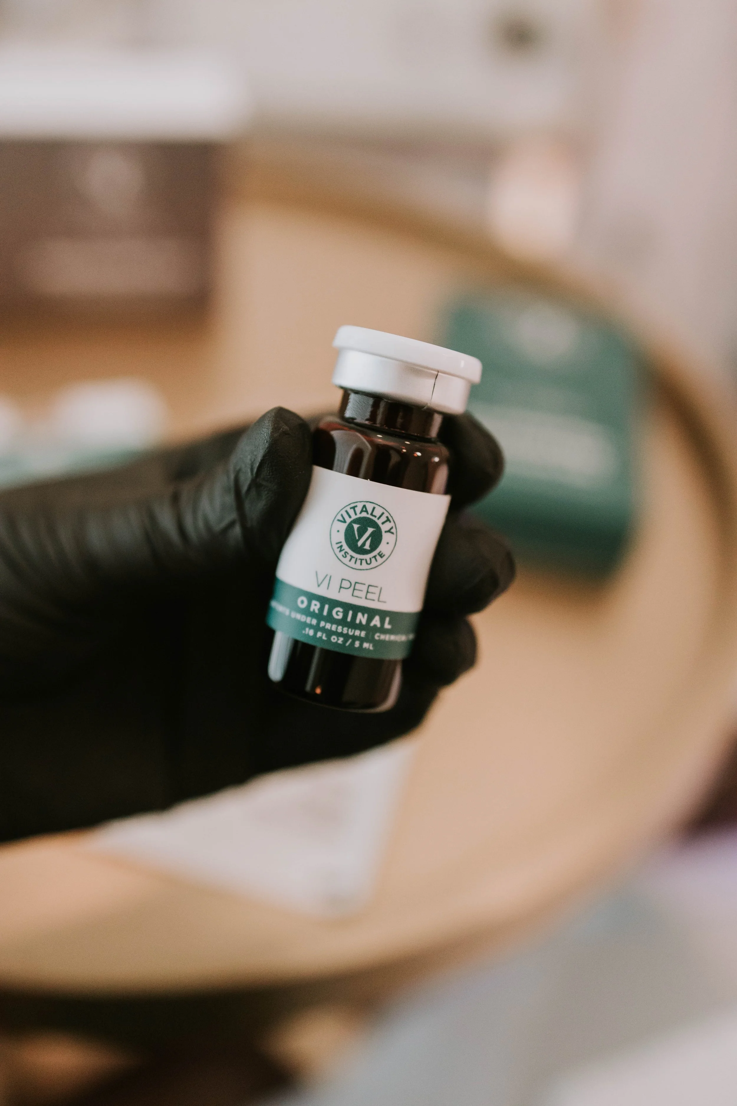 A gloved hand holding a small dark bottle with a white cap, labeled 'VitaliTY Institute VI Peel Original'.