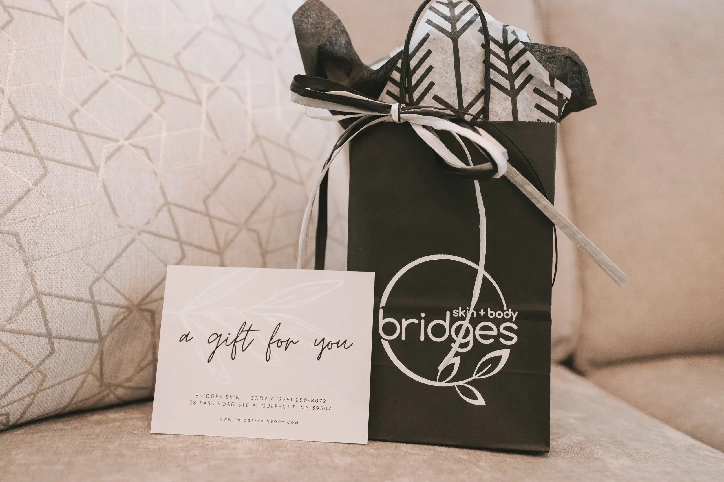 A gift bag from Bridgesskin+body with a black and white patterned wrapping paper and ribbon, sitting next to a gift card on a textured beige surface.