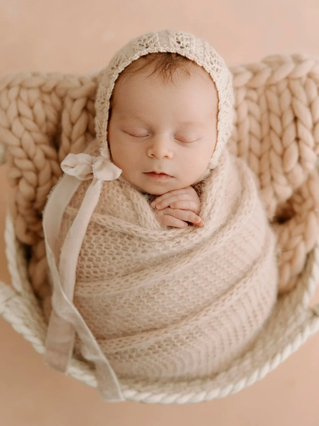 A sleeping baby wrapped in a beige knitted blanket, wearing a matching bonnet, lying in a round woven basket with beige chunky knit pillows.