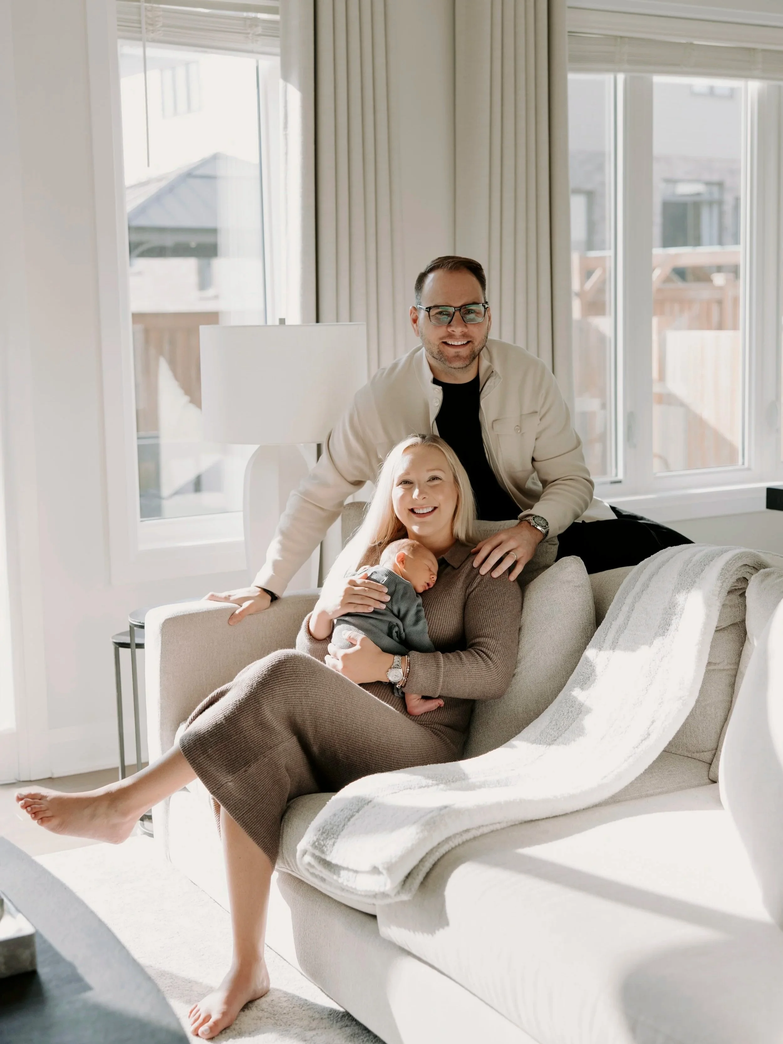 A smiling family of three with light skin in a bright living room, a woman sitting on a sofa holding a newborn baby, a man leaning over smiling, with large windows and curtains in the background.