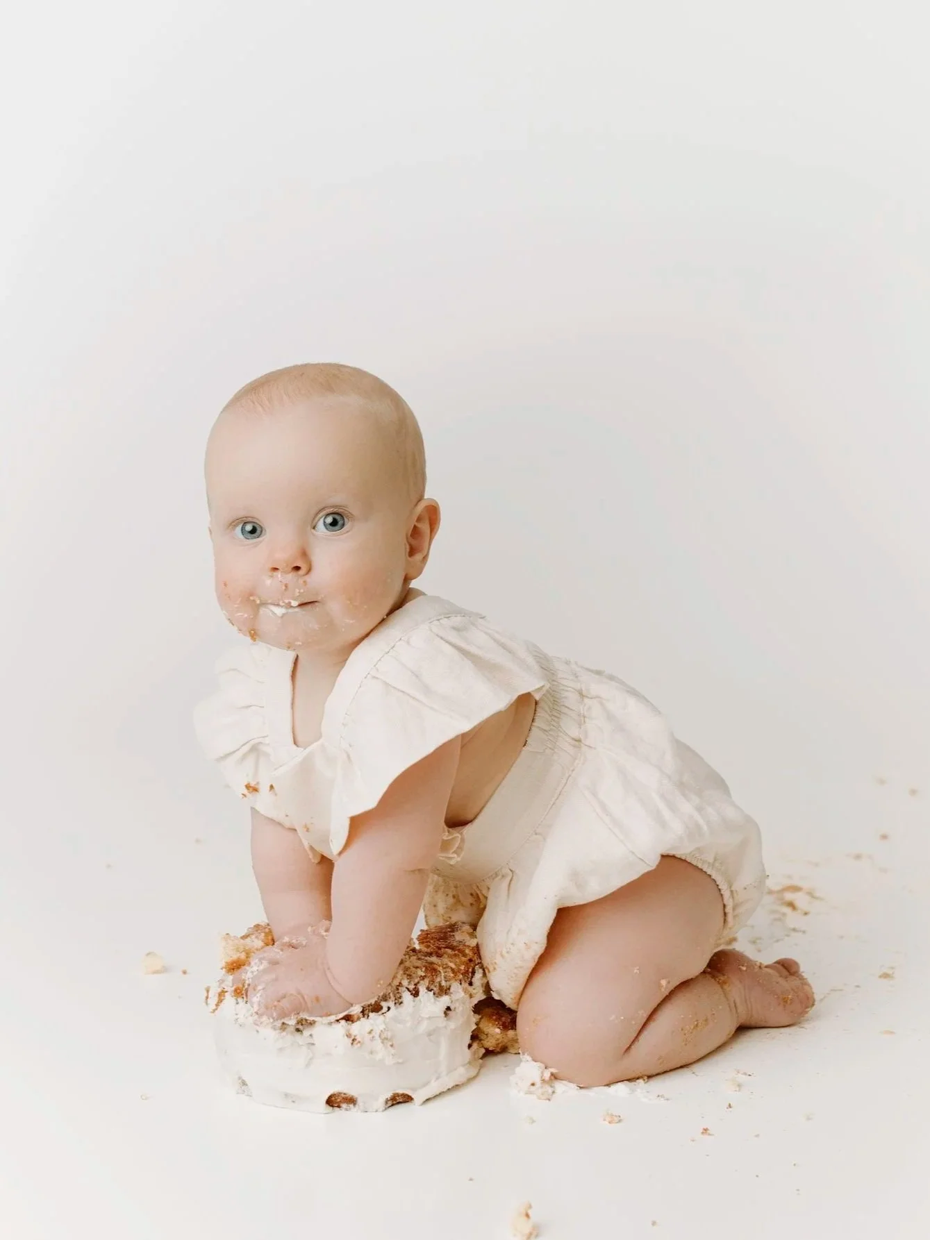 A baby with light skin and blue eyes, wearing a cream-colored dress, is kneeling on a white surface with a piece of cake, with some cake crumbs and frosting on their face and hands, against a plain white background.