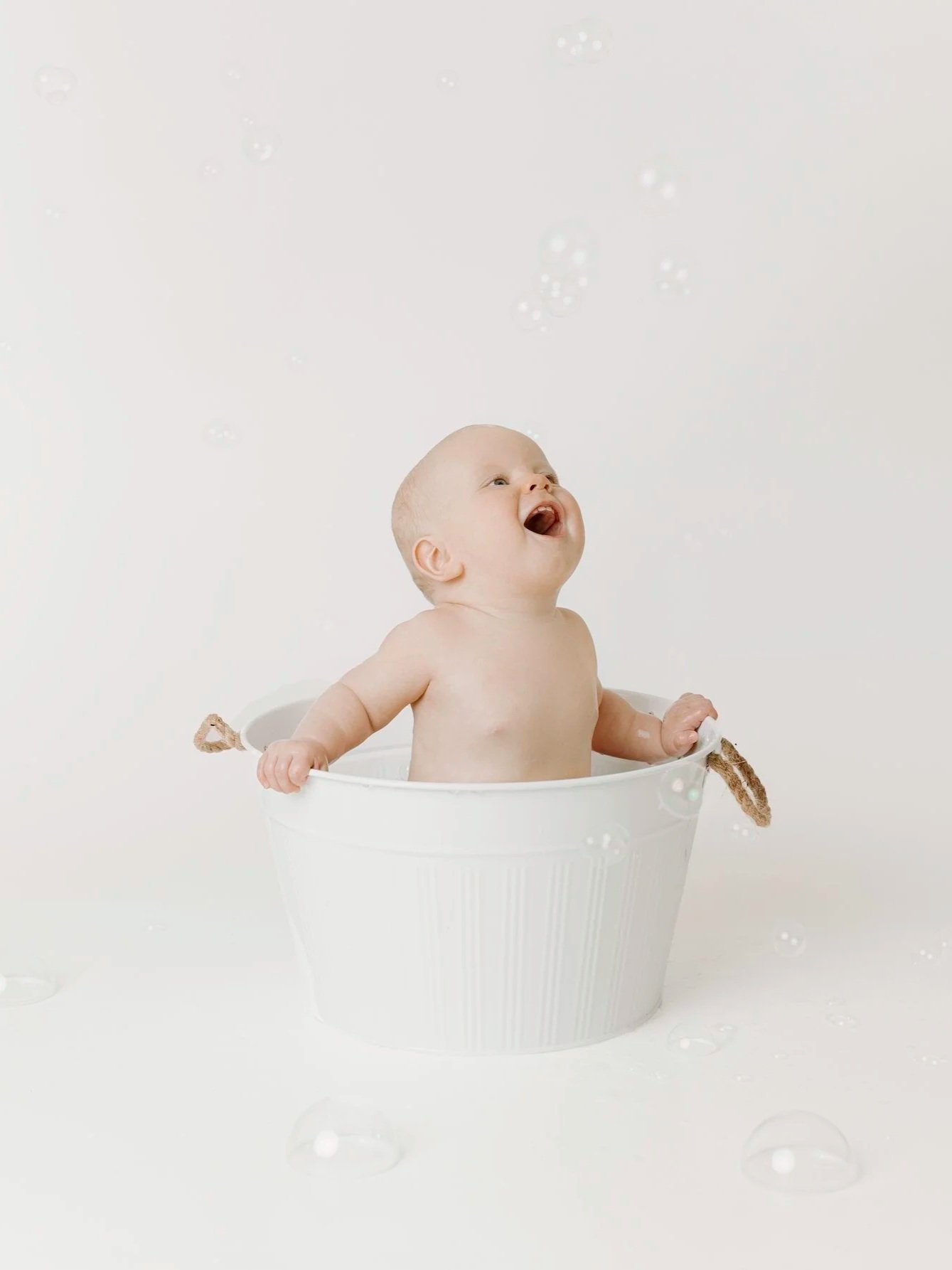 A smiling baby sitting in a white bucket with bubbles floating around on a white background.
