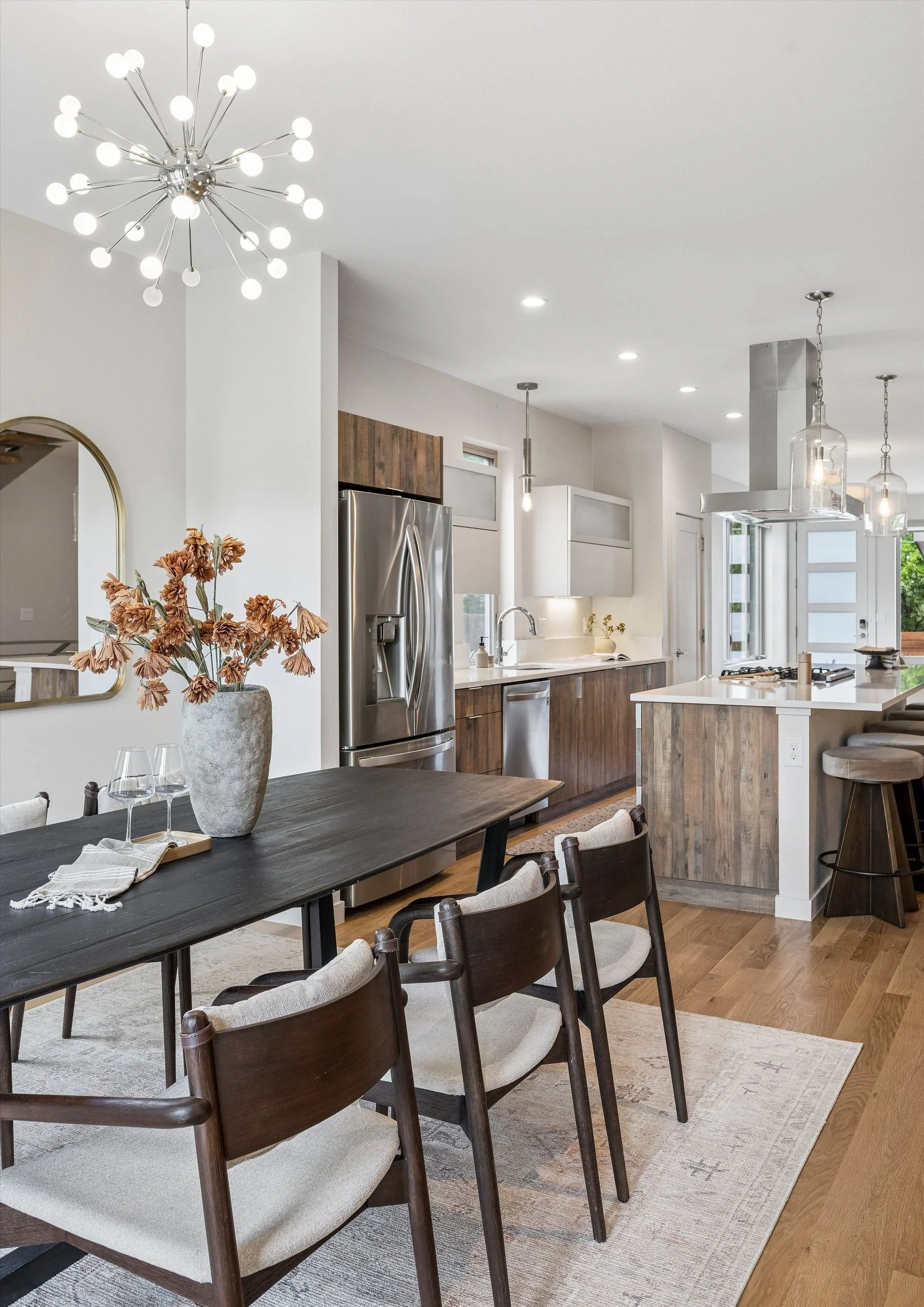 Modern kitchen with wooden cabinets, stainless steel refrigerator, and white countertop. There is a dining table with chairs, a vase with dried flowers, and hanging pendant lights.