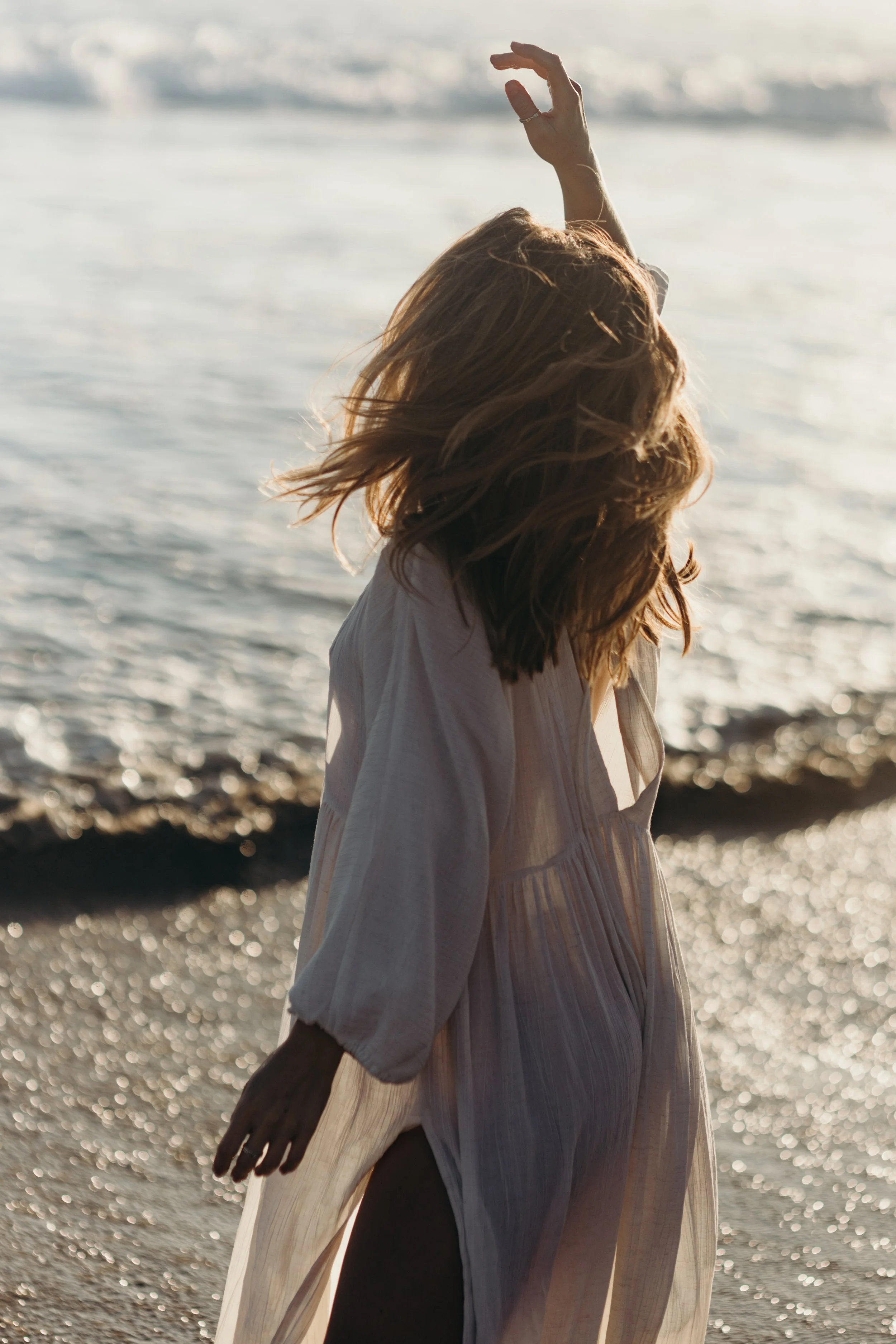 A woman with shoulder-length hair standing by the ocean, with her arm raised, wearing a white dress, during sunset.