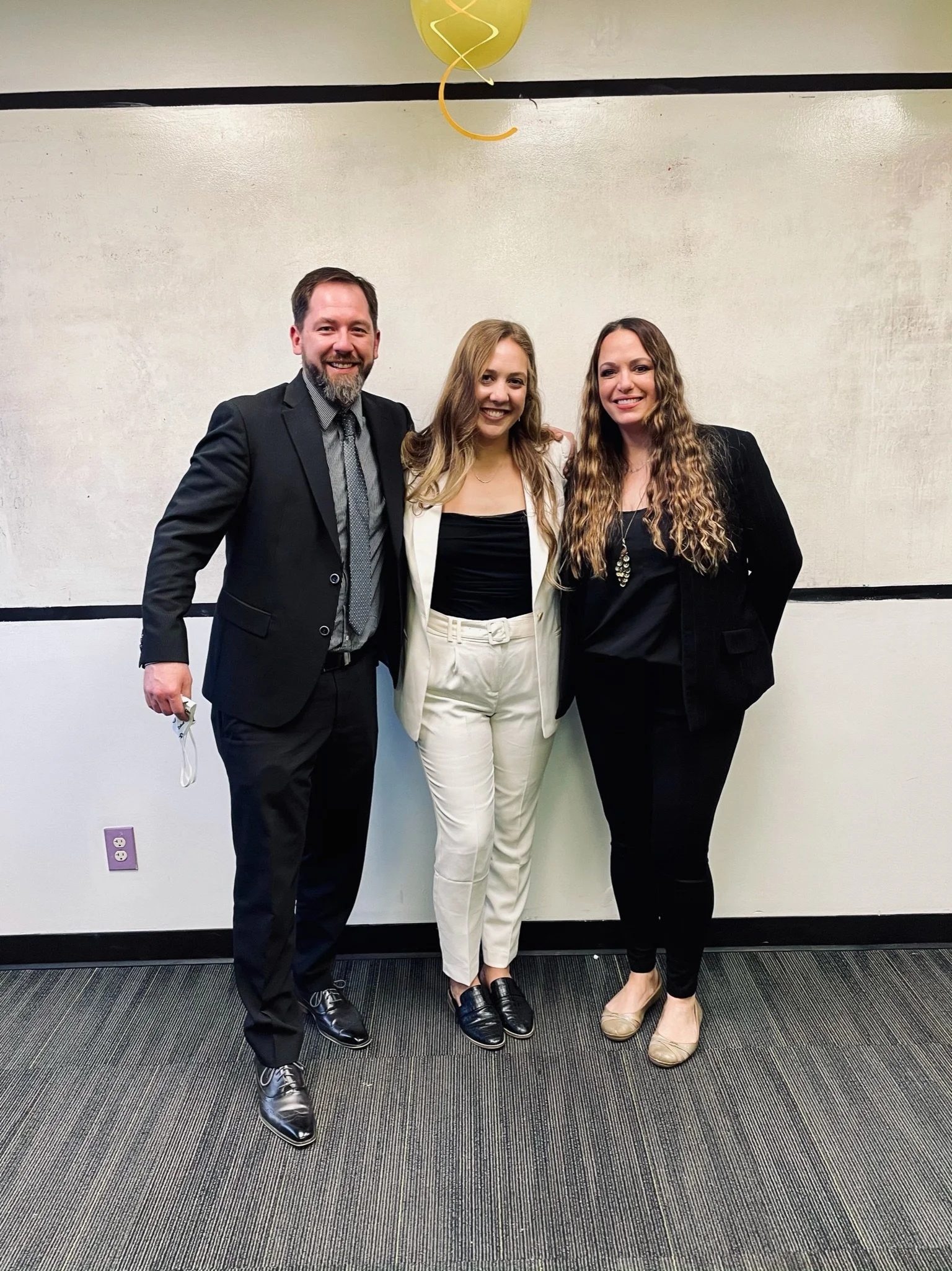 Three people, two women and one man, standing close together and smiling in an indoor setting. They are dressed in formal attire, with the man wearing a suit and the women wearing blazers and dress shoes.