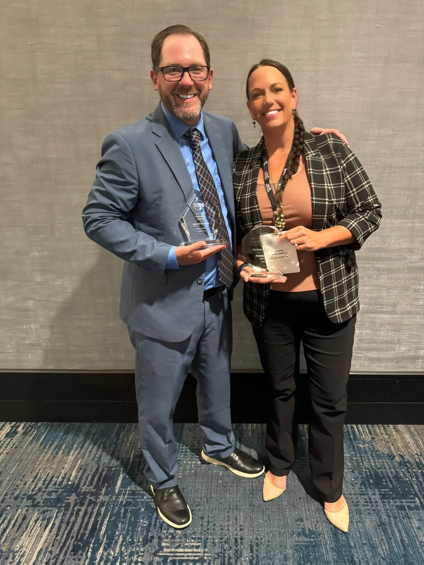 A man and woman dressed in business attire are smiling and holding awards at an indoor event.