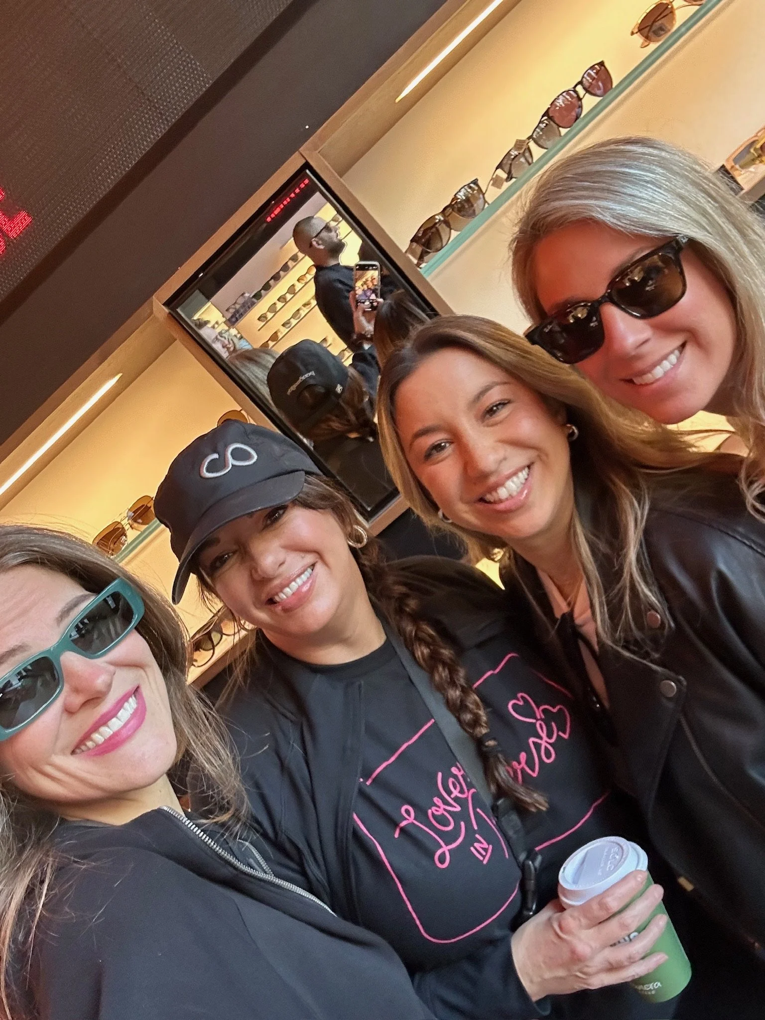 Four women taking a selfie together inside an eyewear store, with displays of sunglasses in the background and a mirror reflecting the scene.