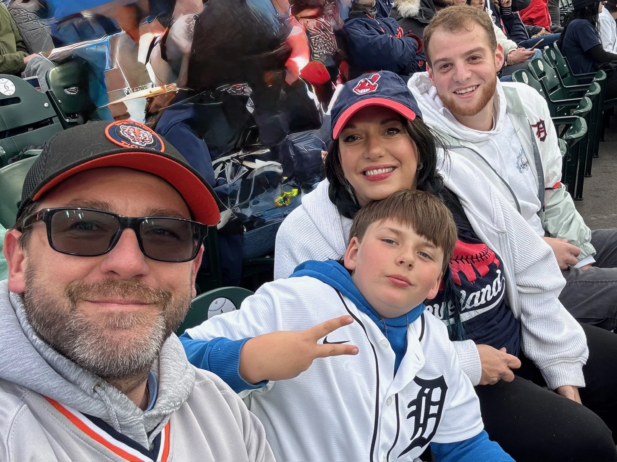 A group of four people sitting in stadium seats at a baseball game, smiling for a selfie. The group includes a man with glasses and a red and black Detroit Tigers cap, a woman with dark hair, a young boy making a peace sign, and a young man with ligh
