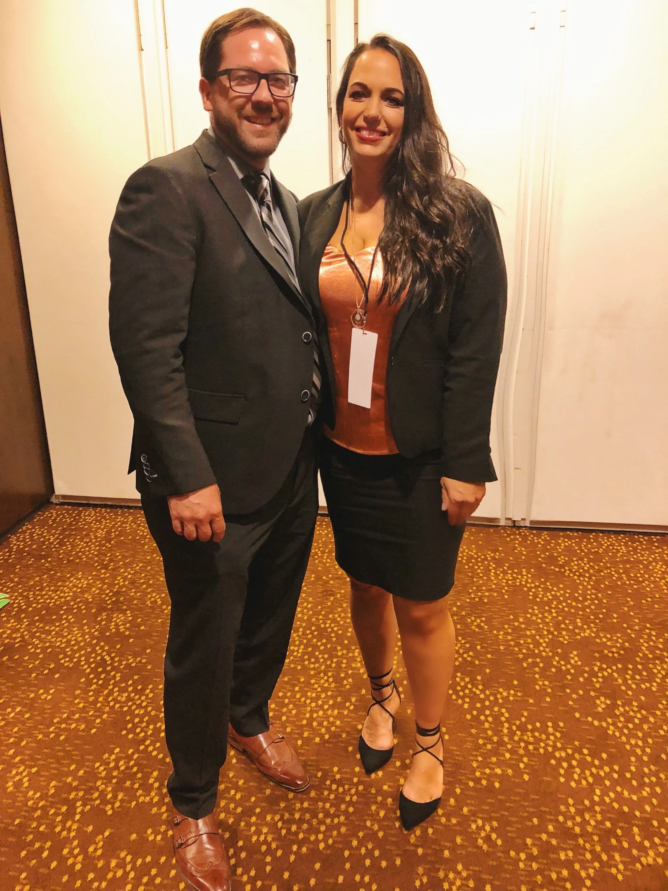 A man and woman dressed in formal attire standing together inside a room, smiling at the camera.