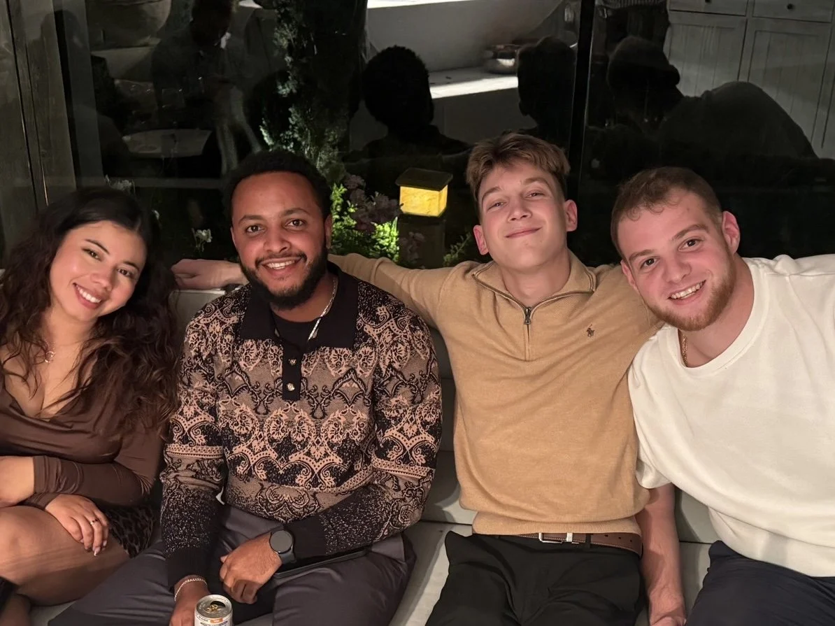 Four young adults sitting close together on a couch, smiling, with their arms around each other, in an indoor setting with a dark, reflective background.