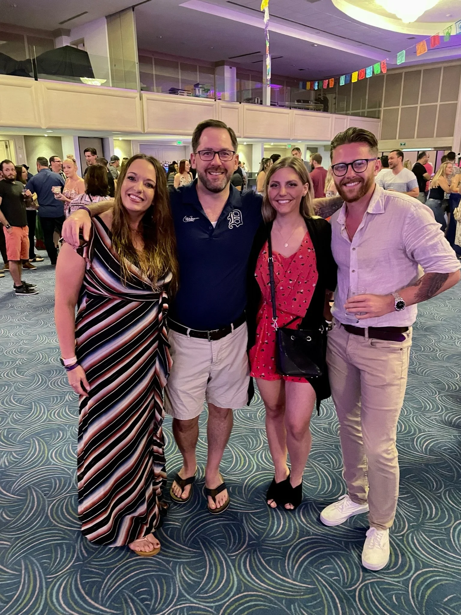 Four friends smiling and standing together at a gathering or party, with a crowded room and colorful decorations in the background.