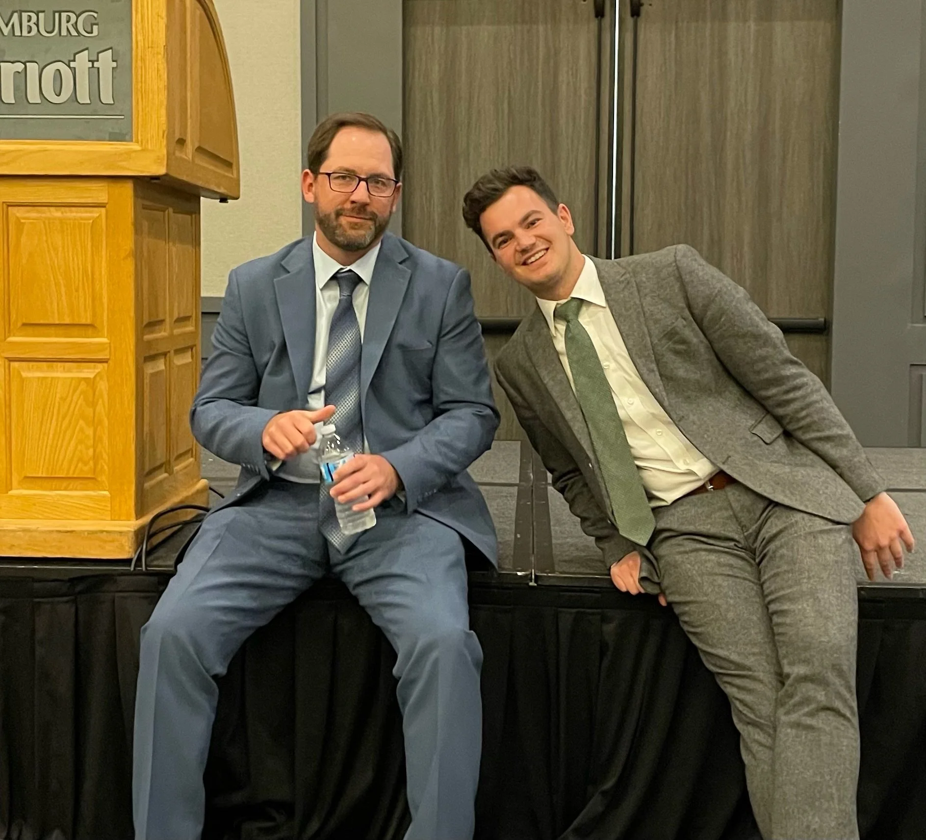 Two men in suits sitting on a stage, one holding a water bottle. Both smiling and leaning slightly towards each other.