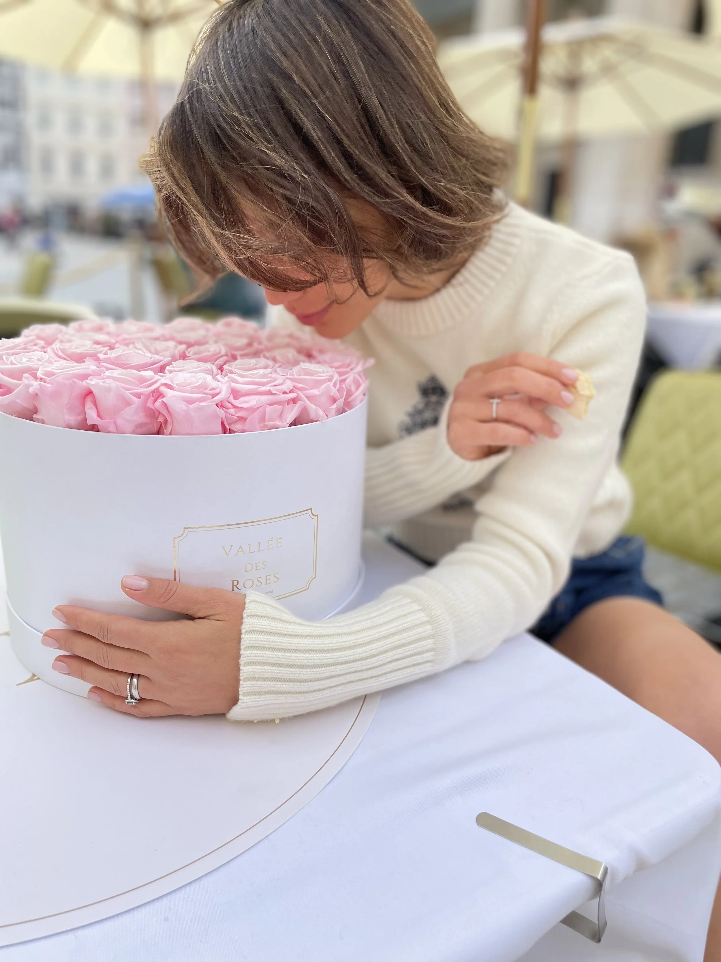 A woman with short brown hair wearing a white sweater and ring, leaning over a large white round box filled with pink roses, on a white table outside at a festive event.