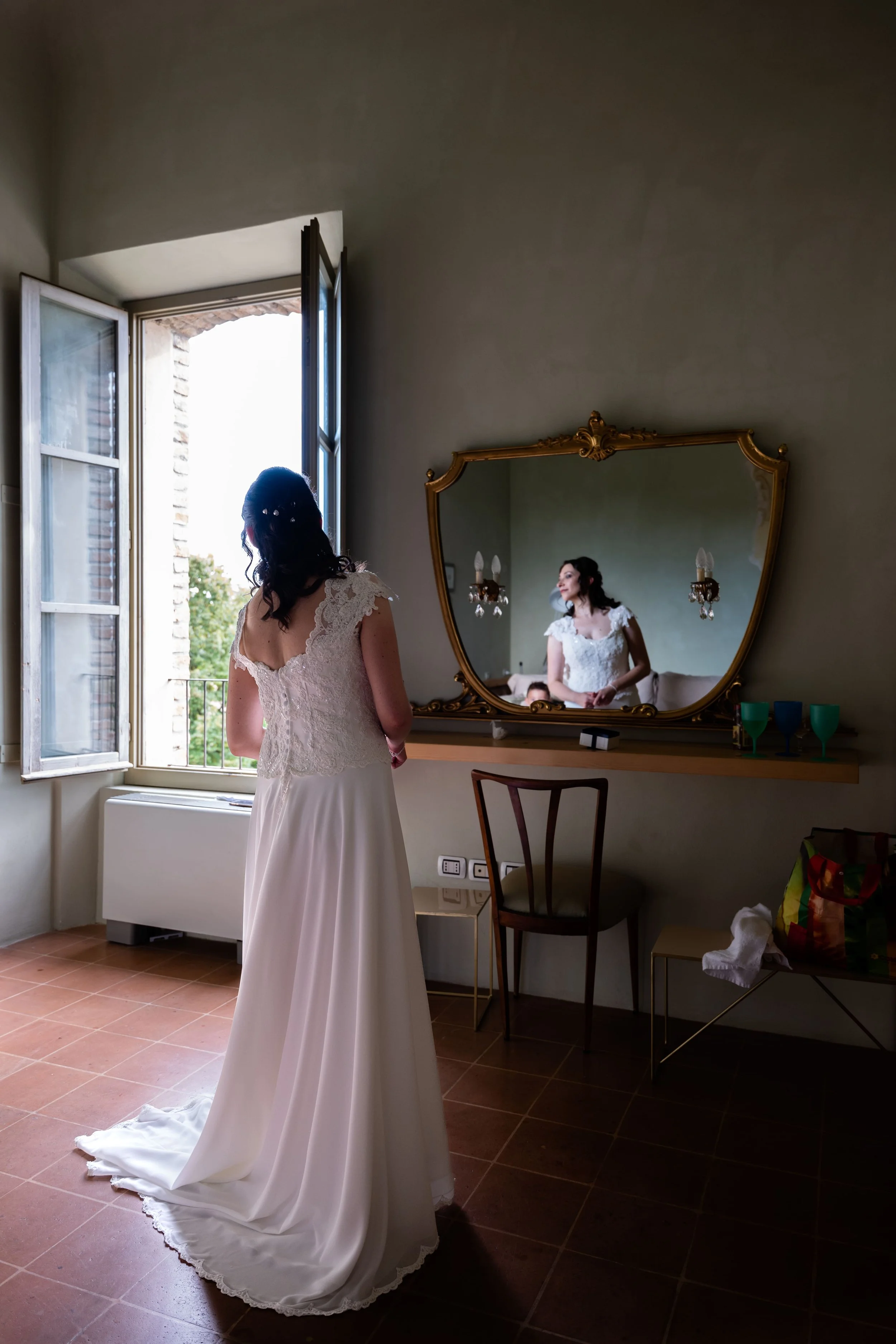 Una donna in un vestito da sposa si guarda nello specchio in una stanza illuminata dalla luce naturale che entra da una finestra aperta.