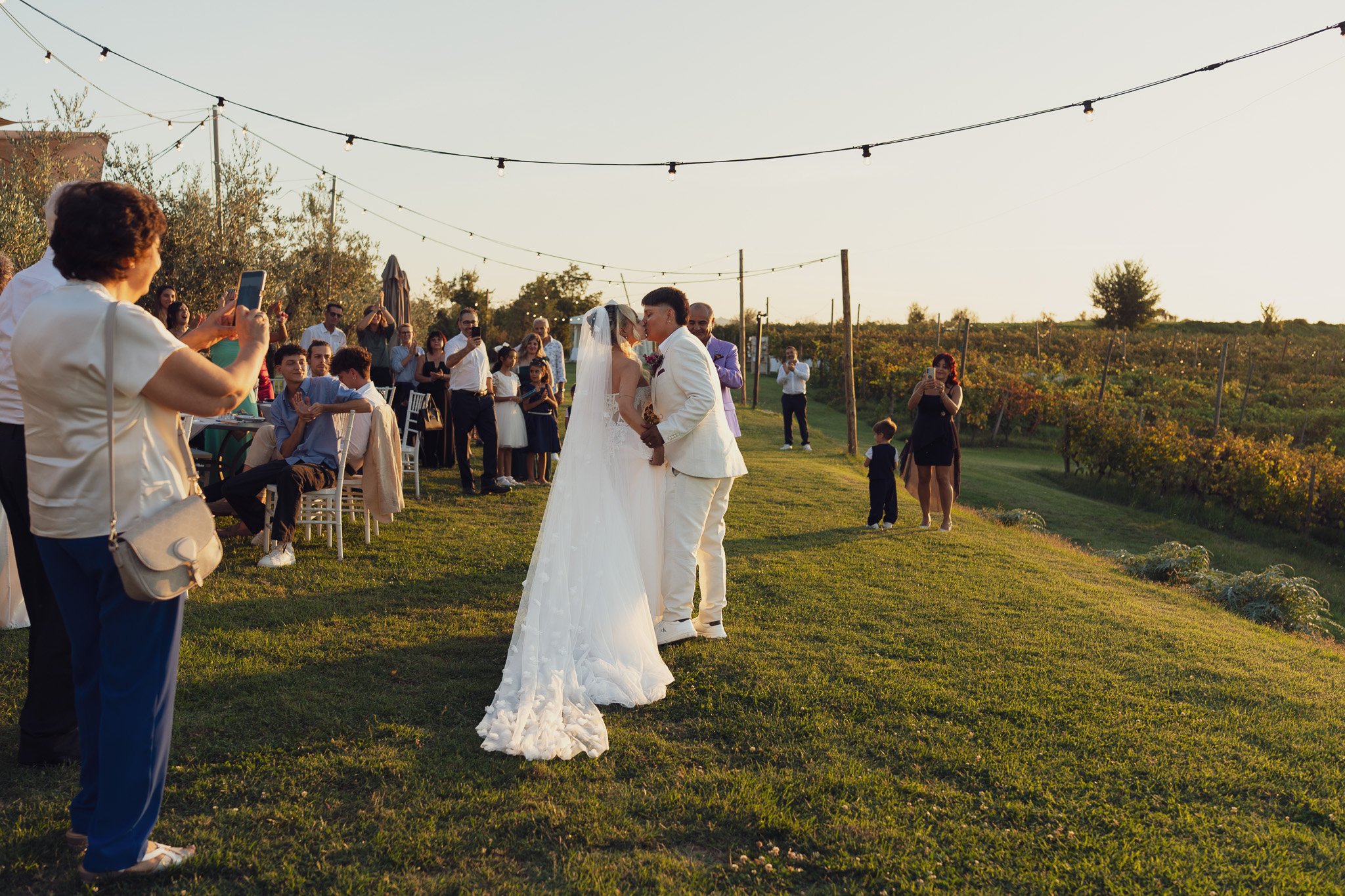 Una coppia di sposi si bacia durante un matrimonio all'aperto al tramonto, circondati da amici e parenti, con luci decorative sospese sopra il prato.