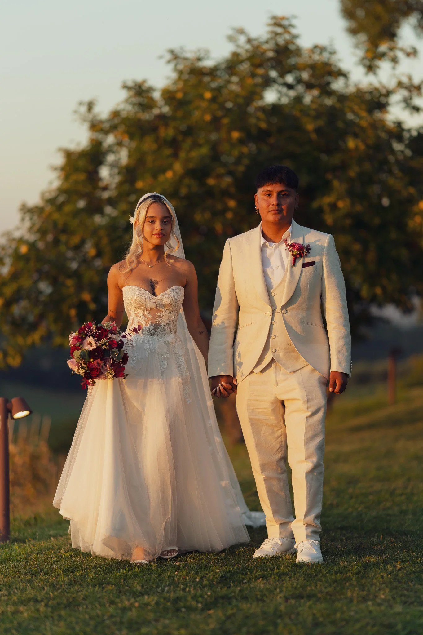 Due persone, una donna e un uomo, vestiti con abiti da sposa e sposo, che si tengono per mano all'aperto al tramonto, con sfondo di alberi e cielo.