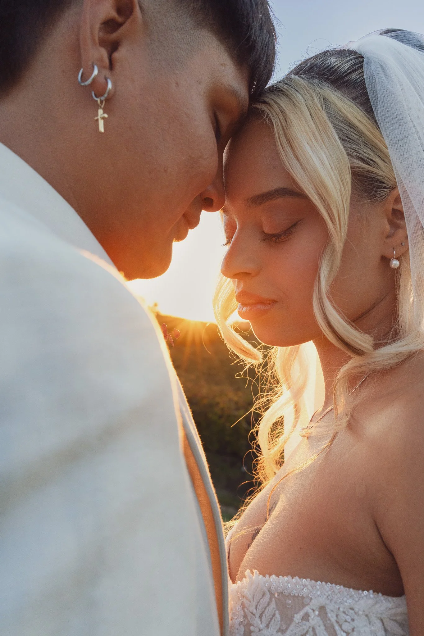 Una coppia di sposi si bacia sotto il sole al tramonto, con abbigliamento da matrimonio, in un ambiente naturale.