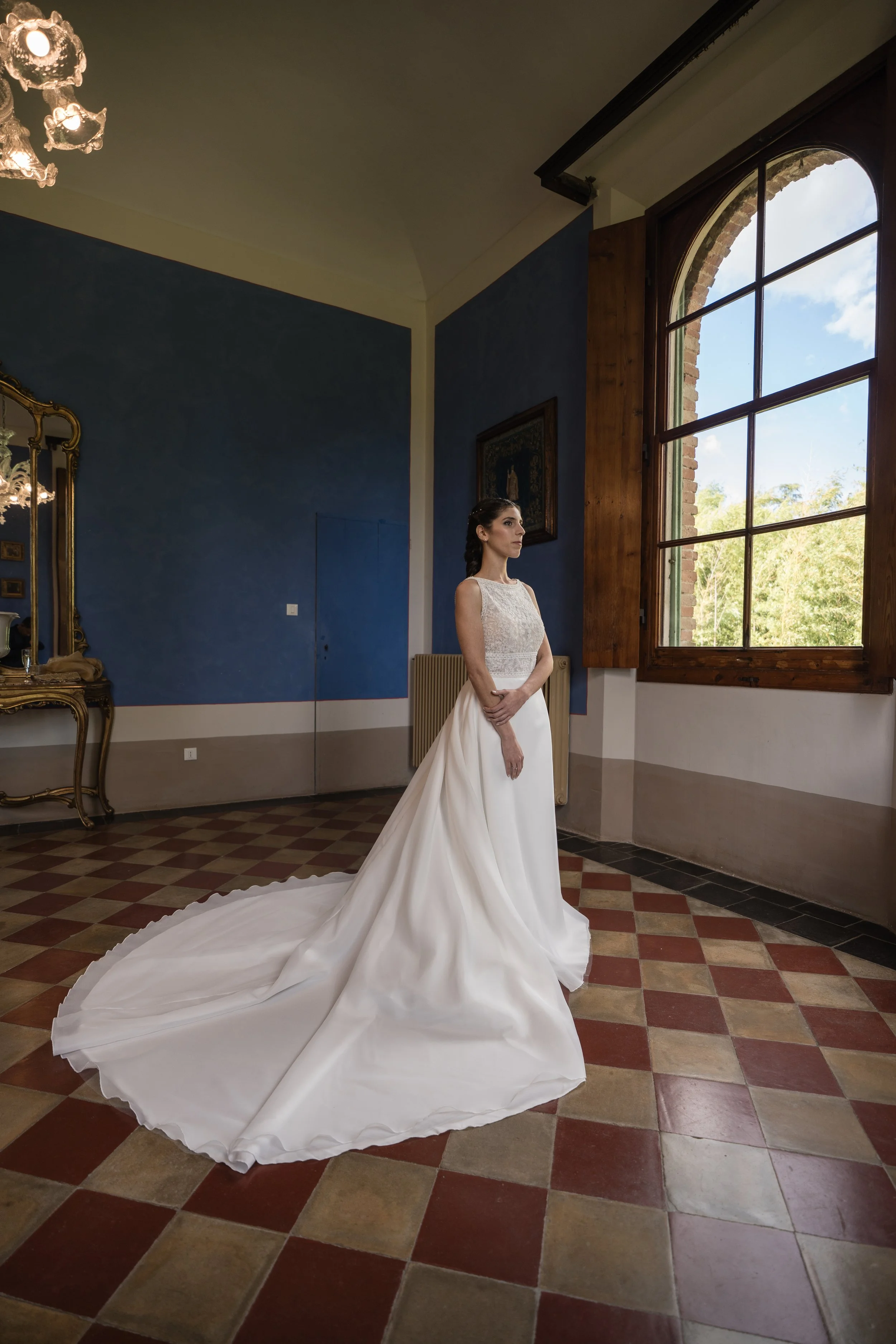 Una donna in abito da sposa bianco all'interno di una stanza con pareti blu e un grande finestrone in legno che mostra un cielo azzurro con nuvole e alberi verdi.
