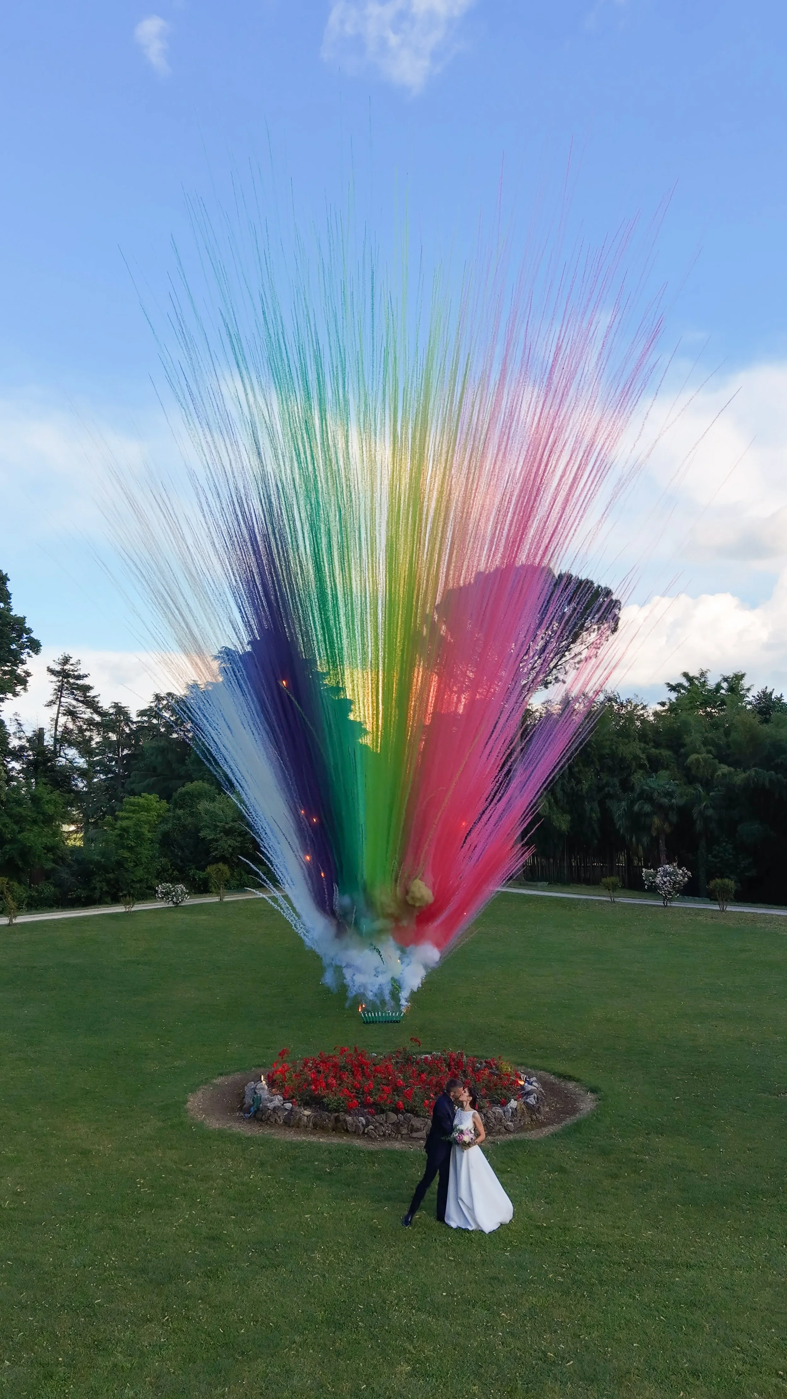 Un fuochista d'acqua colorata spara uno spray di colori sotto un cielo sereno, con un prato verde e alberi sullo sfondo. In primo piano, una coppia di sposini si bacia.