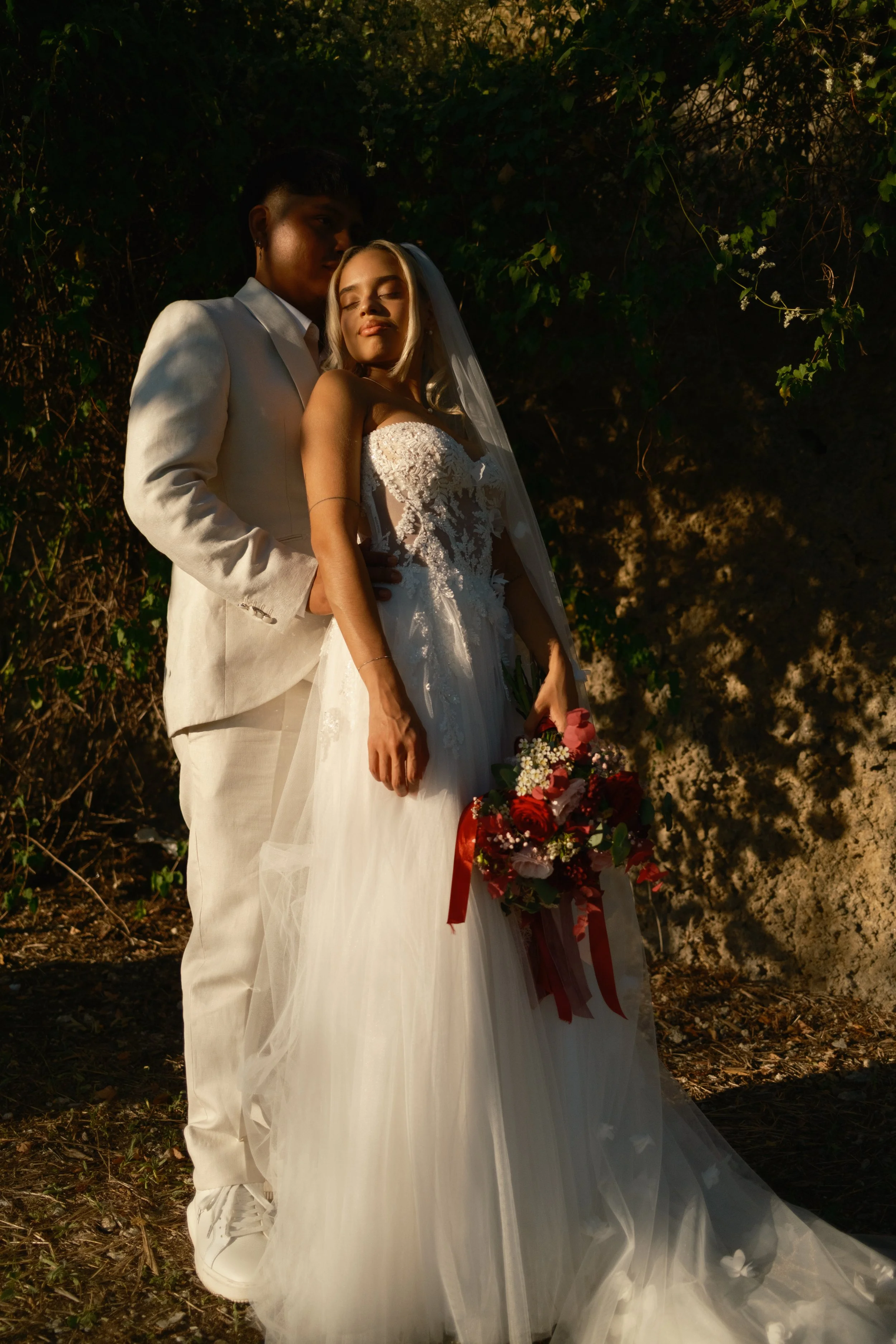 Una coppia di sposi, una donna con abito da sposa e un uomo con completo bianco, si abbracciano all'aperto di sera, con uno sfondo di vegetazione.