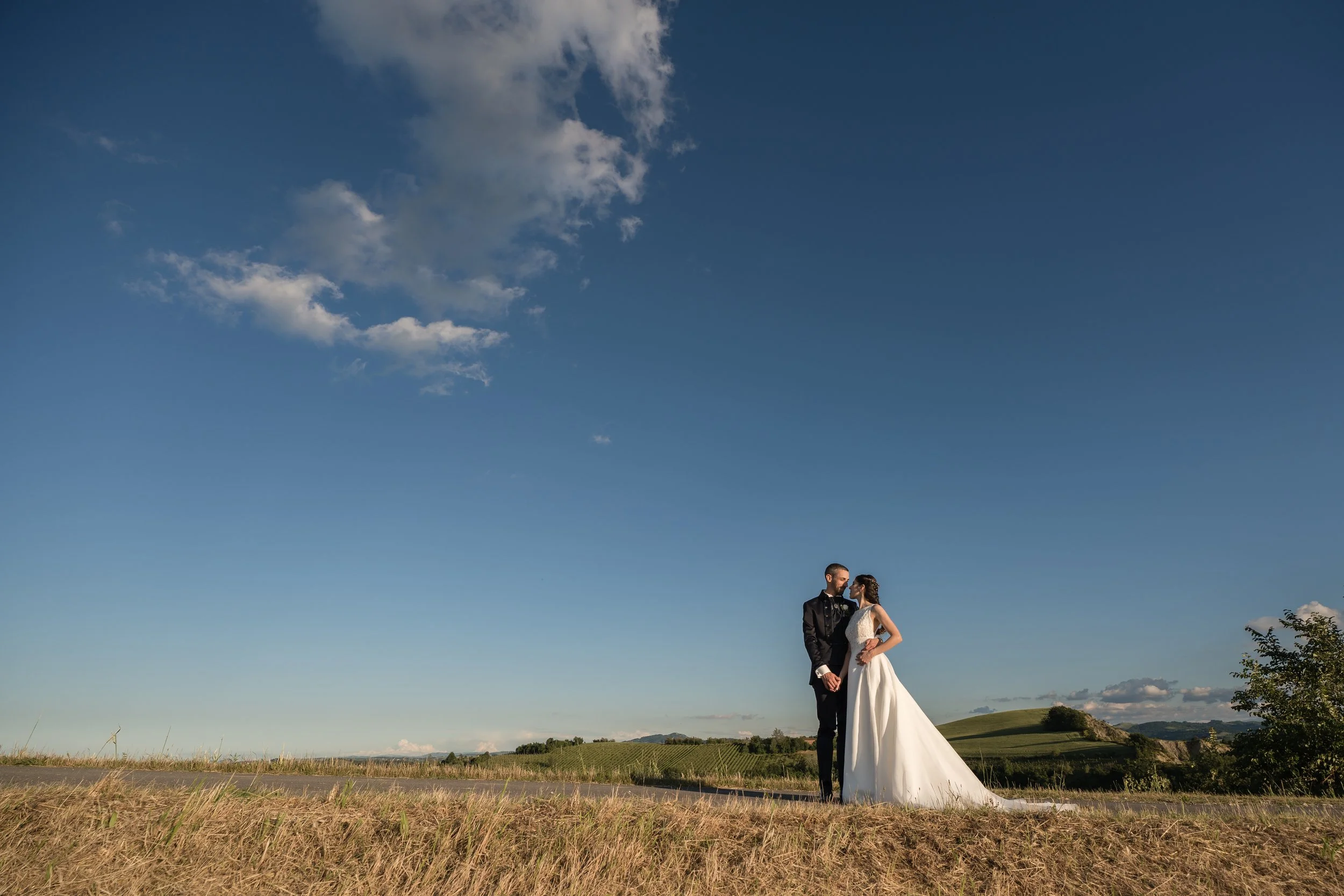 Una coppia in abiti da sposa si tiene per mano in un campo all'aperto sotto un cielo blu con alcune nuvole.