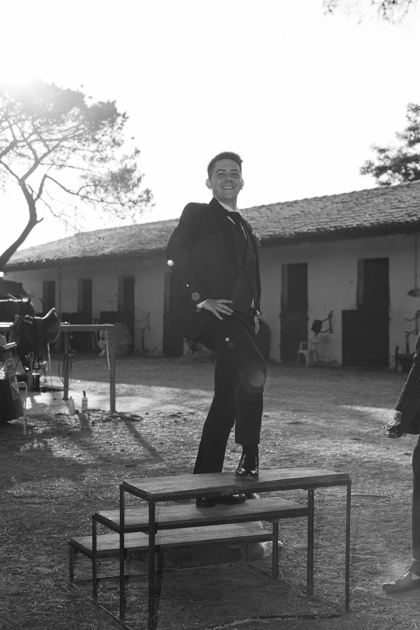 Una persona sorridente in abito formale si trova su una piattaforma di legno sopra una struttura simile a un palco. Sullo sfondo, un maneggio e un albero con rami spogli. L'immagine è in bianco e nero. Marina Romea