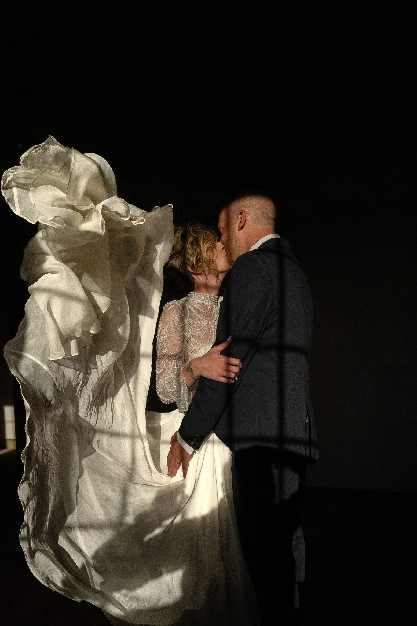 Una coppia si bacia durante il giorno di nozze, con l'abito da sposa e l'abito da sposo, contro uno sfondo scuro.