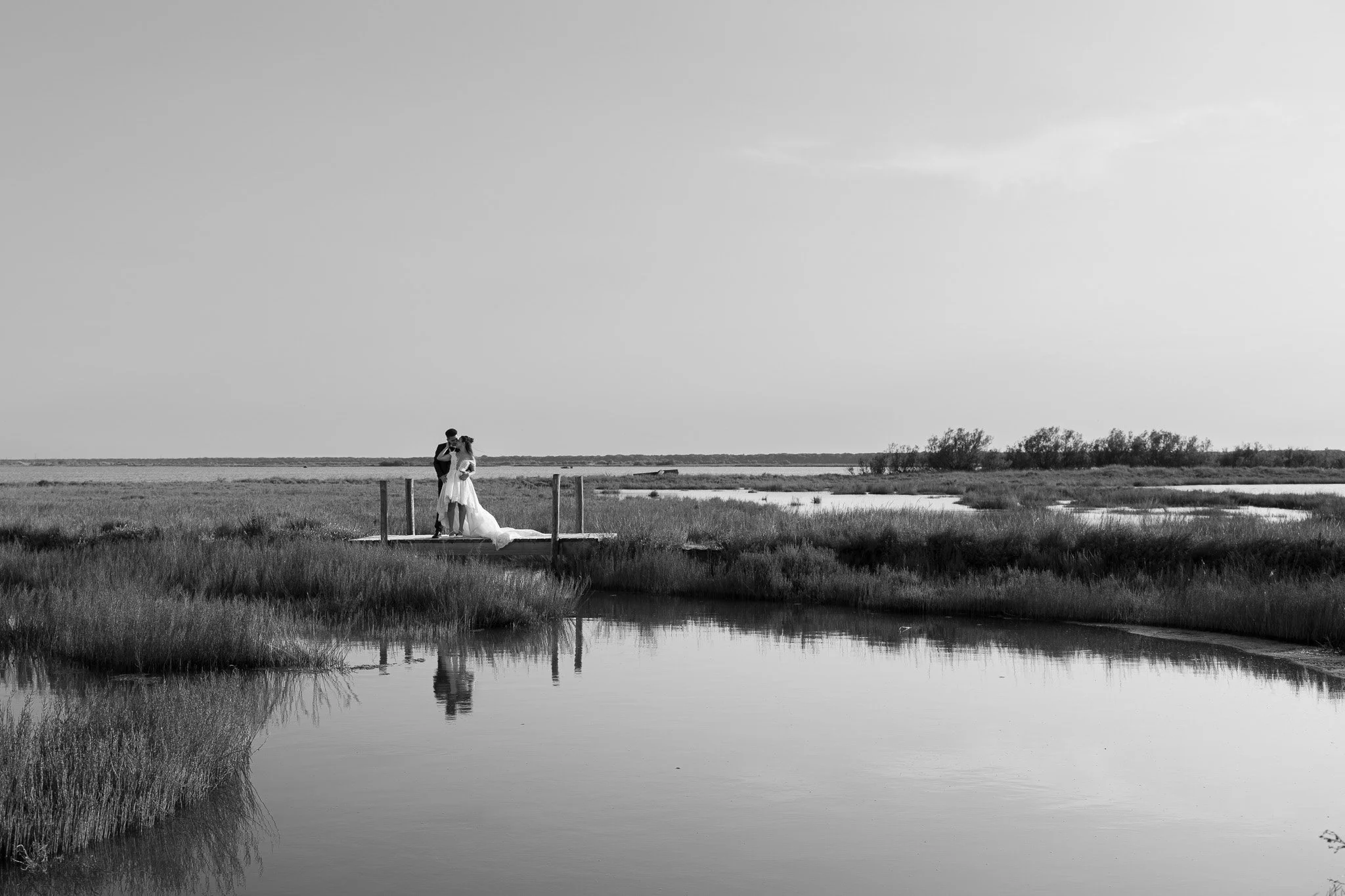Una coppia di sposi si bacia su una piattaforma della Pialassa della Baiona a Ravenna in un paesaggio naturale con vegetazione e un cielo nuvoloso.