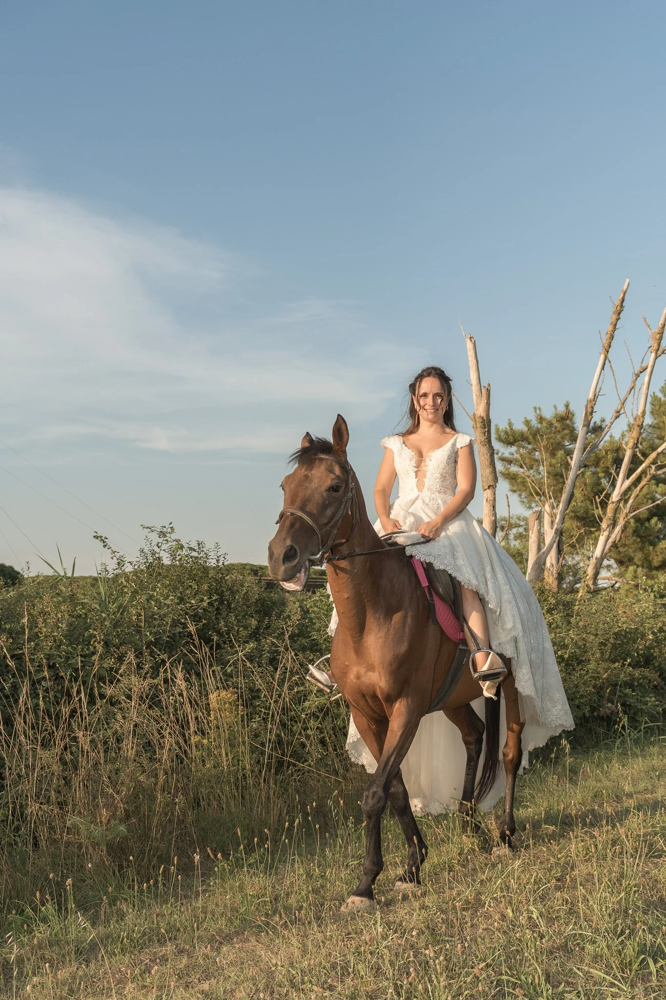 Una donna in abito da sposa seduta su un cavallo in un paesaggio naturale con cielo azzurro e vegetazione. Maneggio di Marina Romea a Ravenna.