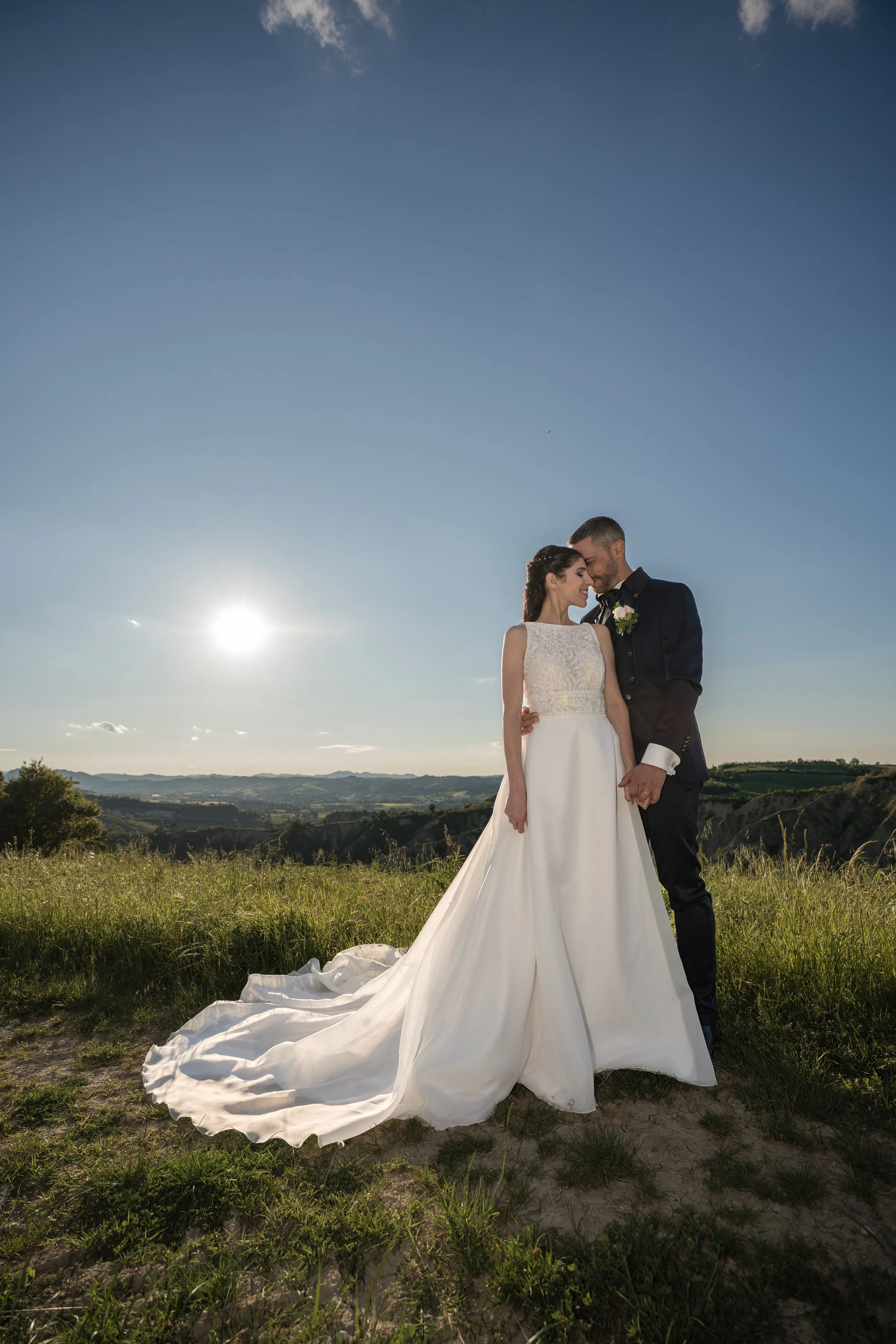 Una coppia di sposi in abiti da matrimonio si tiene per mano in un campo aperto con vista sulle colline, al tramonto o all'alba, sotto un cielo blu con poche nuvole.