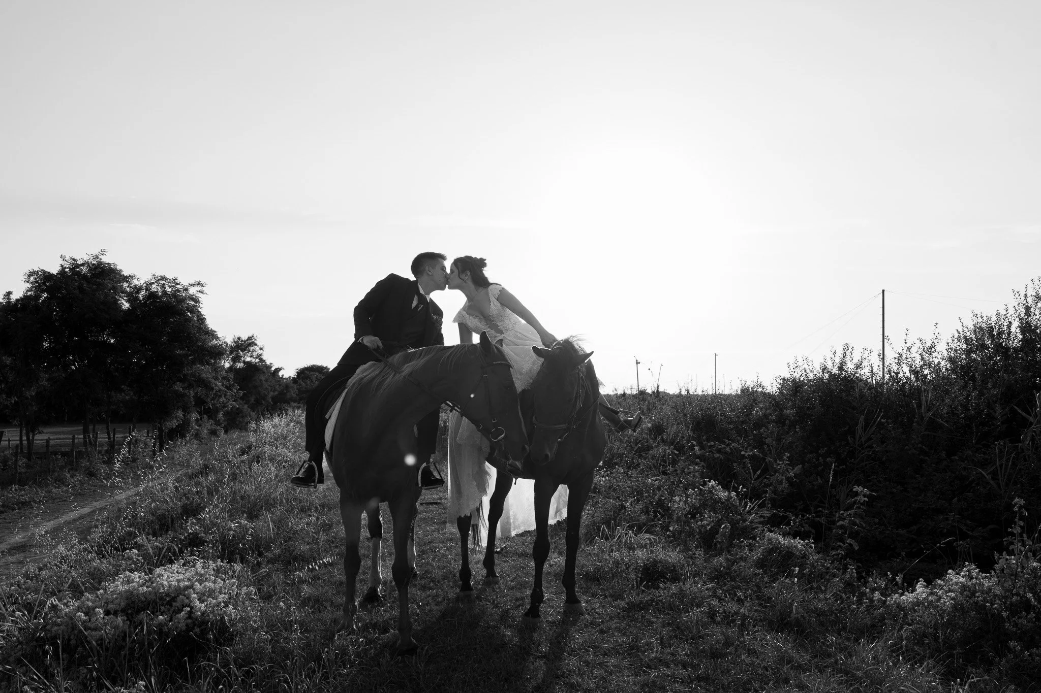 Una coppia di sposi si bacia mentre è seduta su un cavallo, in un paesaggio all'aperto con alberi e cielo al tramonto. Maneggio di Marina Romea a Ravenna.