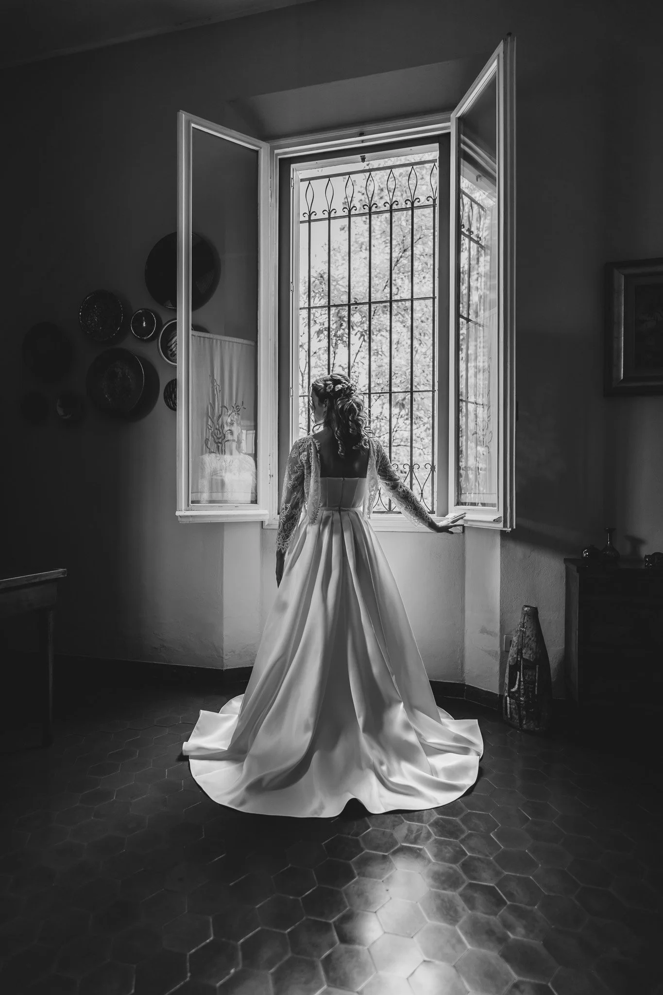 Una donna con un abito da sposa bianco davanti a una finestra aperta, in una stanza con pareti chiare e pavimento in piastrelle esagonali scure, con decorazioni alle pareti e un quadro appeso.