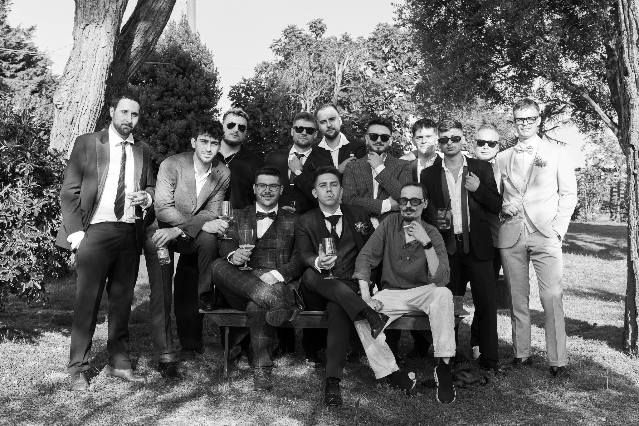 Gruppo di uomini vestiti eleganti con occhiali da sole, alcuni con bicchieri di vino, nel parco della Cascina del Riesling a Ravenna, in un contesto di Matrimonio.