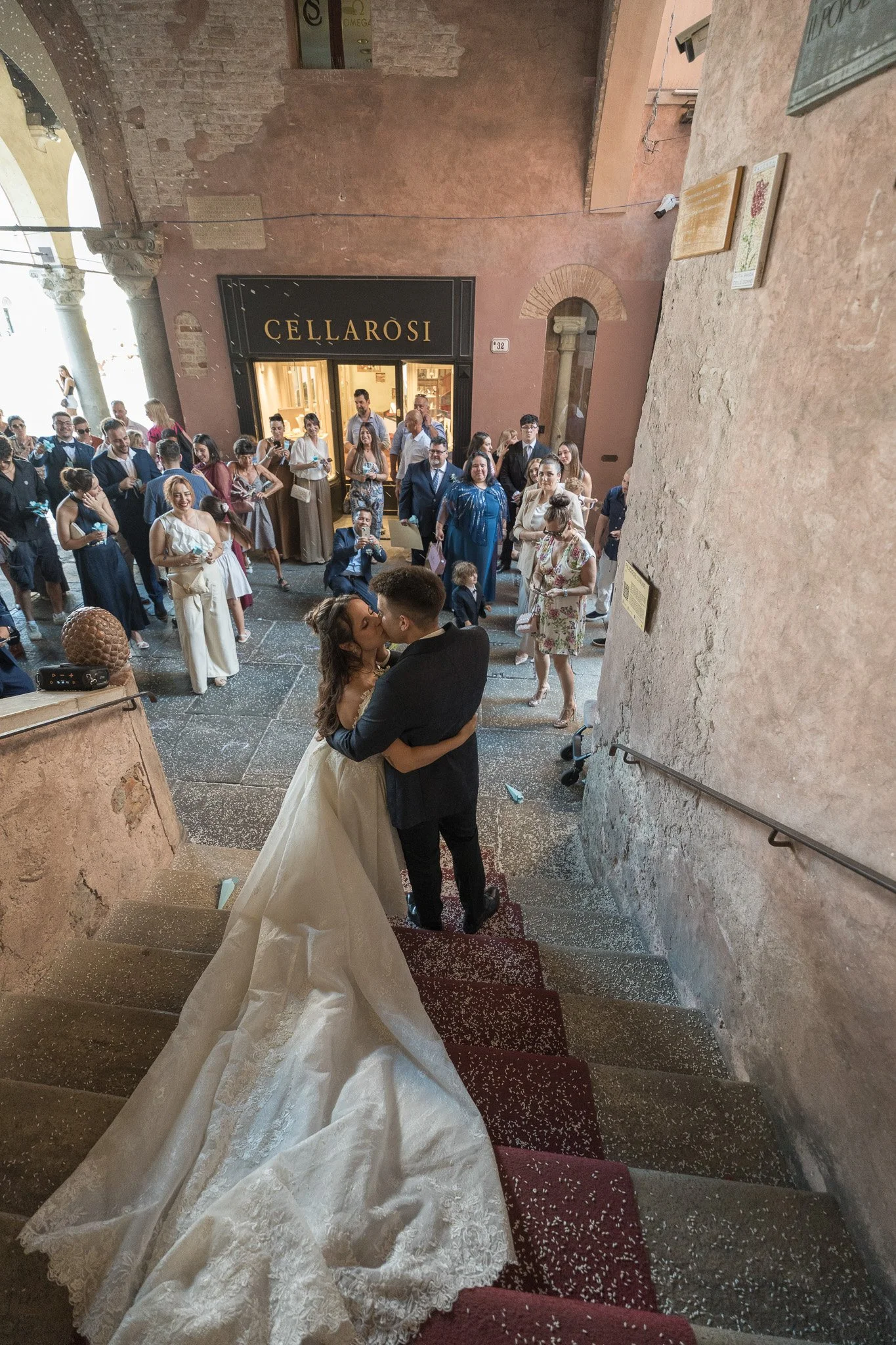 Un matrimonio si bacia sulla scala di pietra durante il matrimonio in Comune a Ravenna, con molti invitati che osservano all'ingresso di un edificio antico con pareti in pietra e decorazioni.
