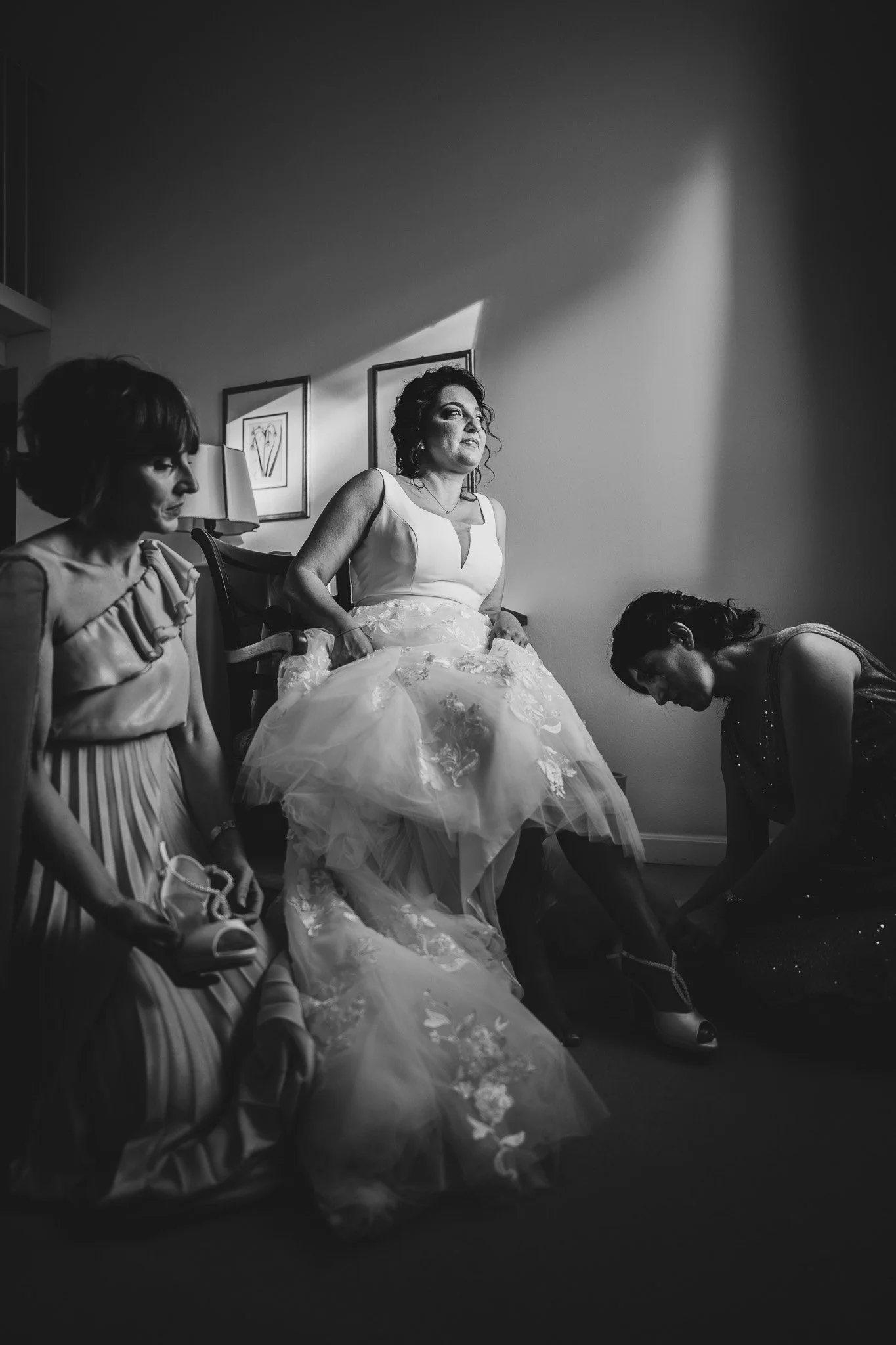 Un gruppo di donne, una delle quali indossa un abito da sposa, si sta preparando probabilmente per un matrimonio. La scena è in una stanza con pareti chiare e ha decorazioni di quadri alle pareti.