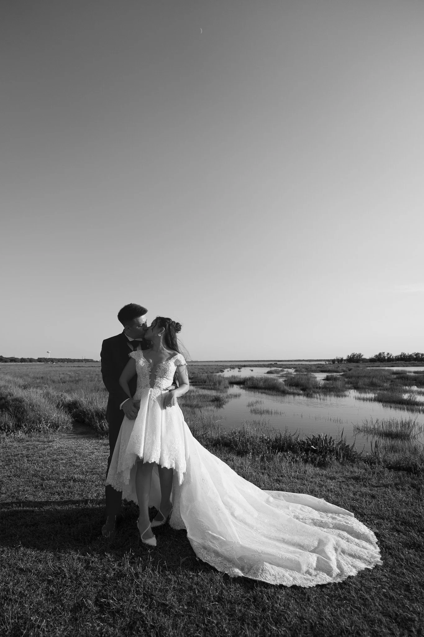 Una coppia di sposi si bacia all'aperto, con uno sfondo di campagna e un lago, in bianco e nero. Il luogo è a Marina Romea di Ravenna.