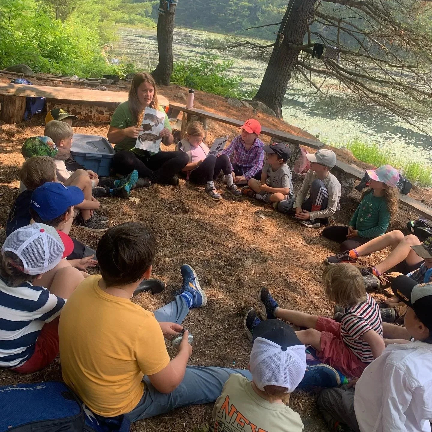 A group of children and an adult woman sitting in a semicircle on the ground outdoors near a lake, listening to the woman who is holding pictures of fish.