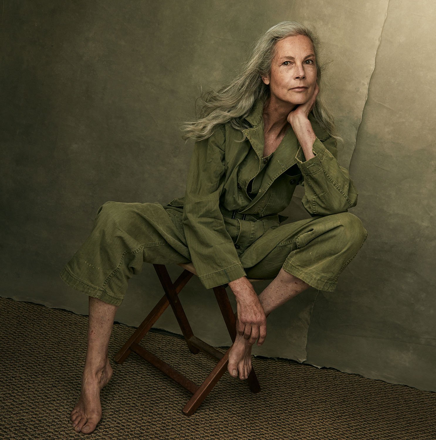 Elegant barefoot woman with long gray hair in a green jumpsuit, seated in Aaron Jay Young’s Los Angeles portrait studio against a textured backdrop, exuding natural beauty and quiet confidence.