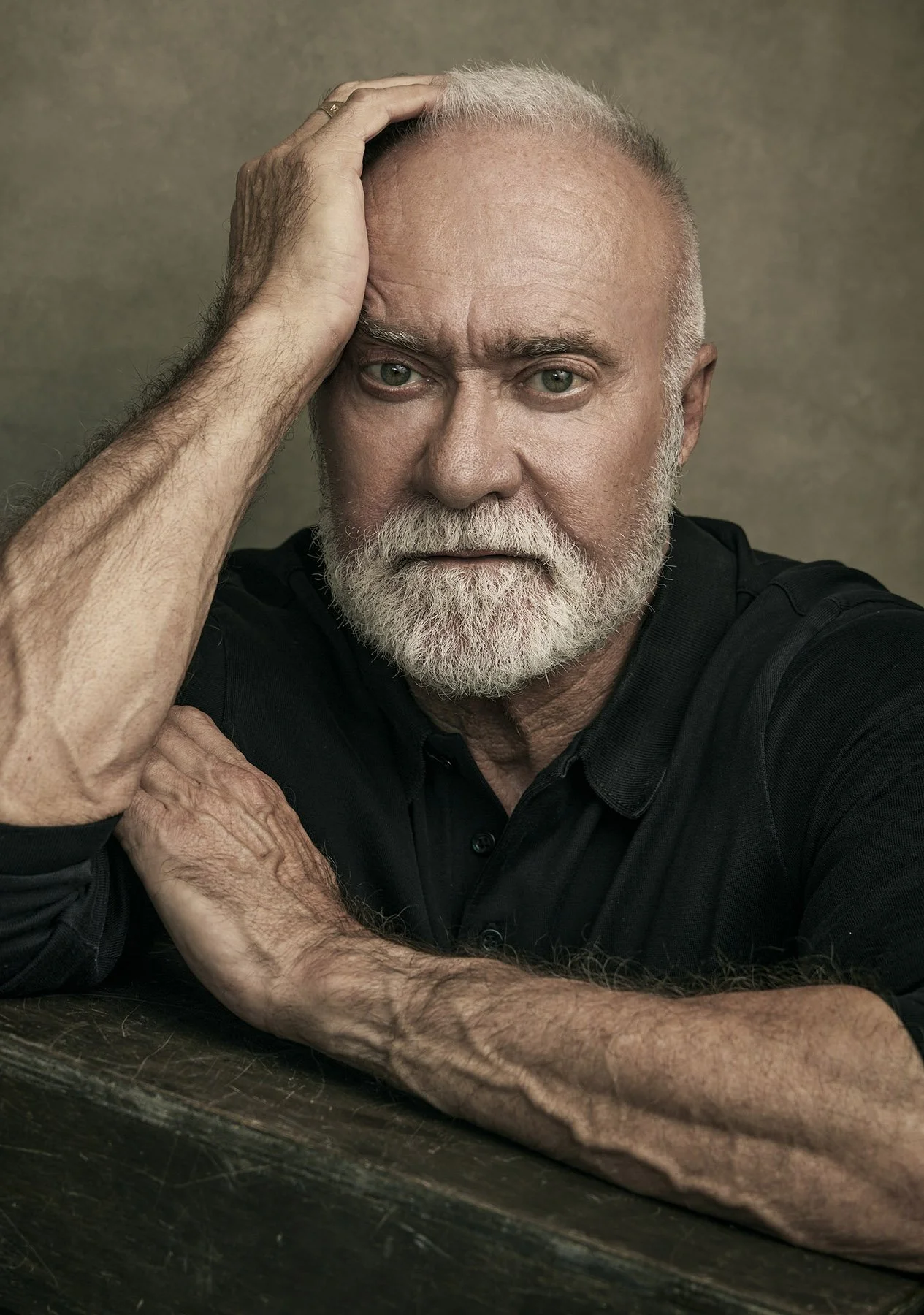 Color portrait of a man resting his head on his hand, looking into the lens with intensity and depth, against a warm-toned backdrop.