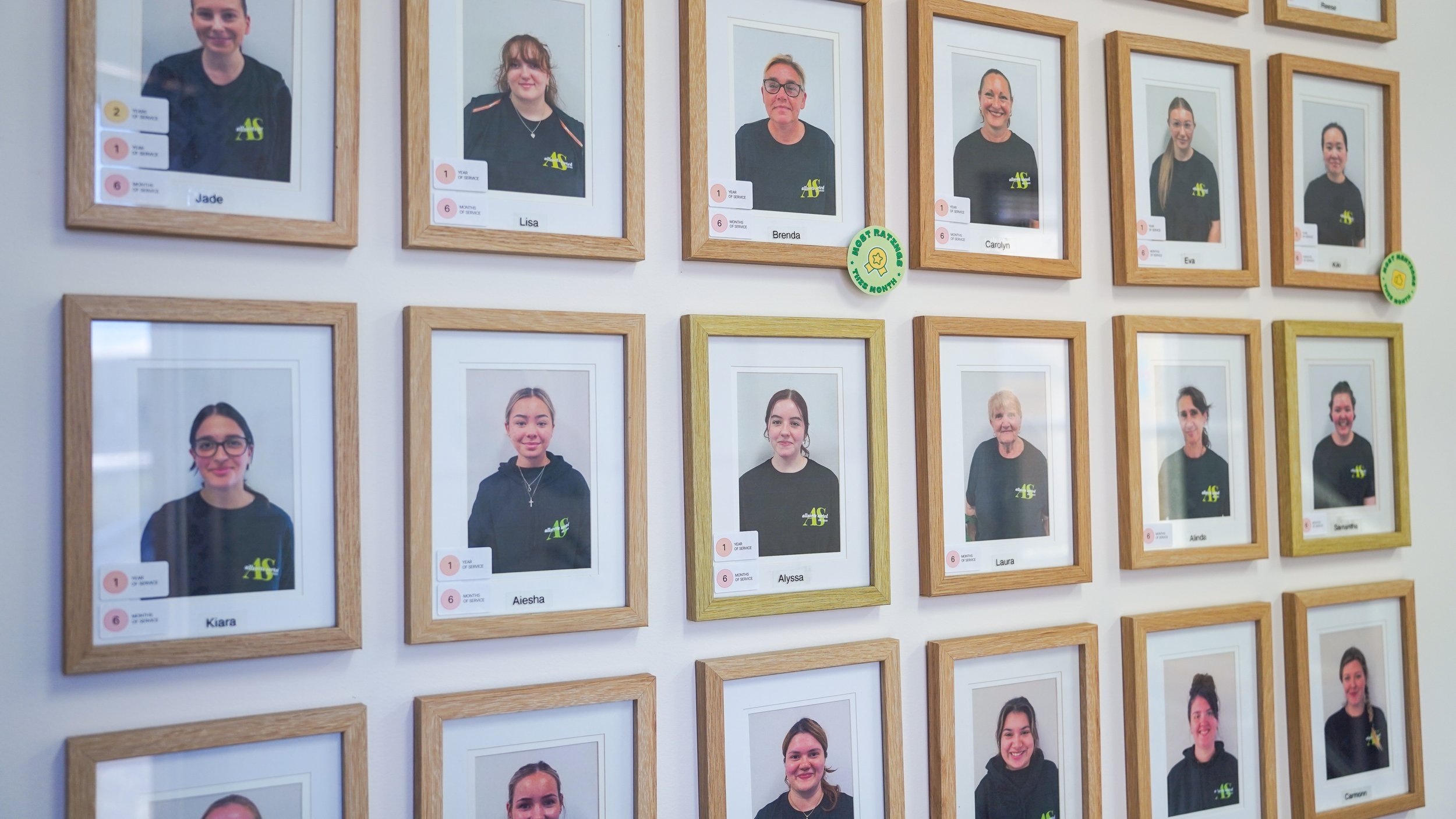 Wall displaying framed photographs of individuals, with name tags and some green circular stickers indicating recognition or awards.