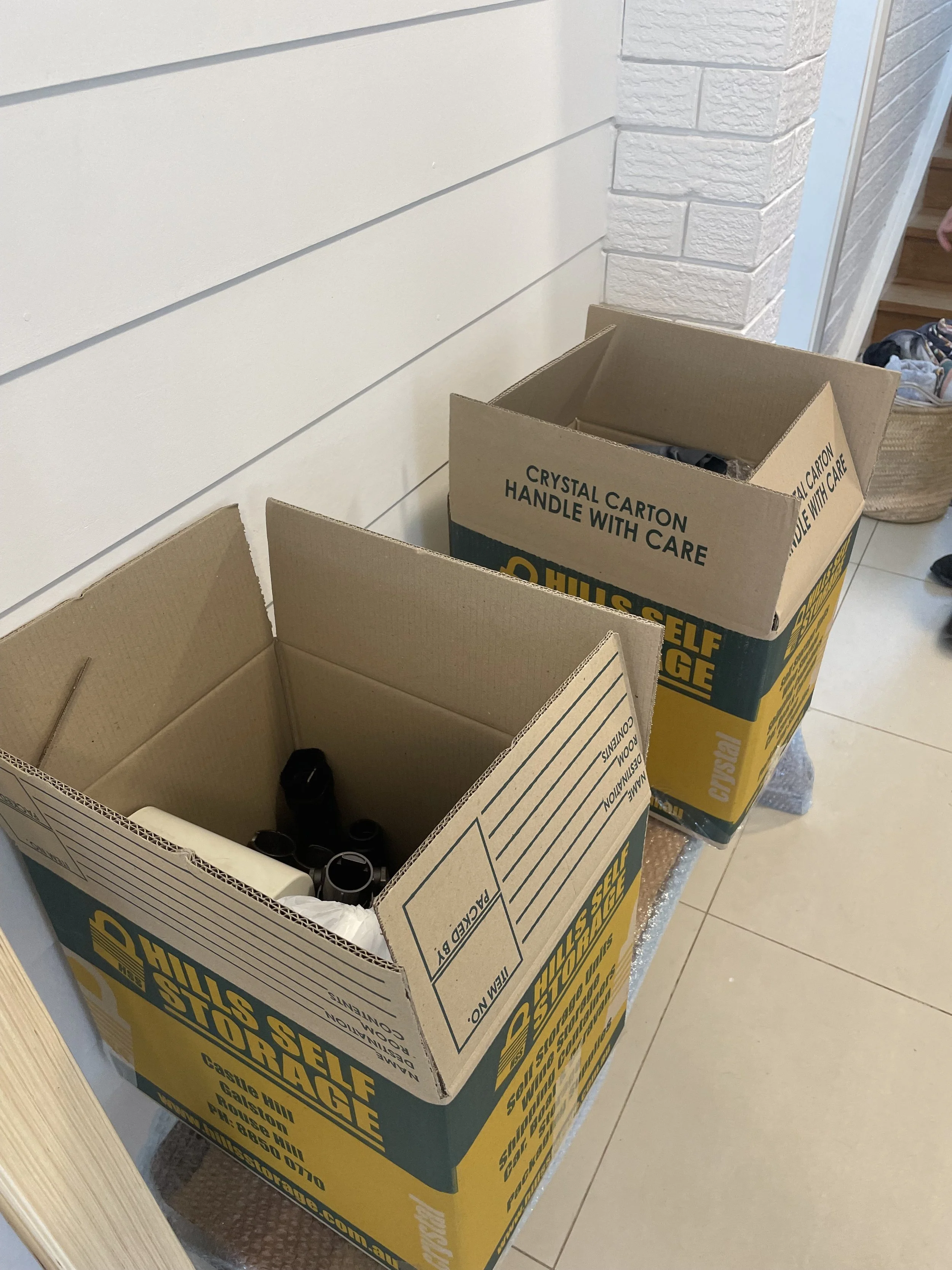 Two open cardboard boxes labeled 'Hills Self Storage' containing items wrapped in bubble wrap, situated on a tiled floor next to a white wall.
