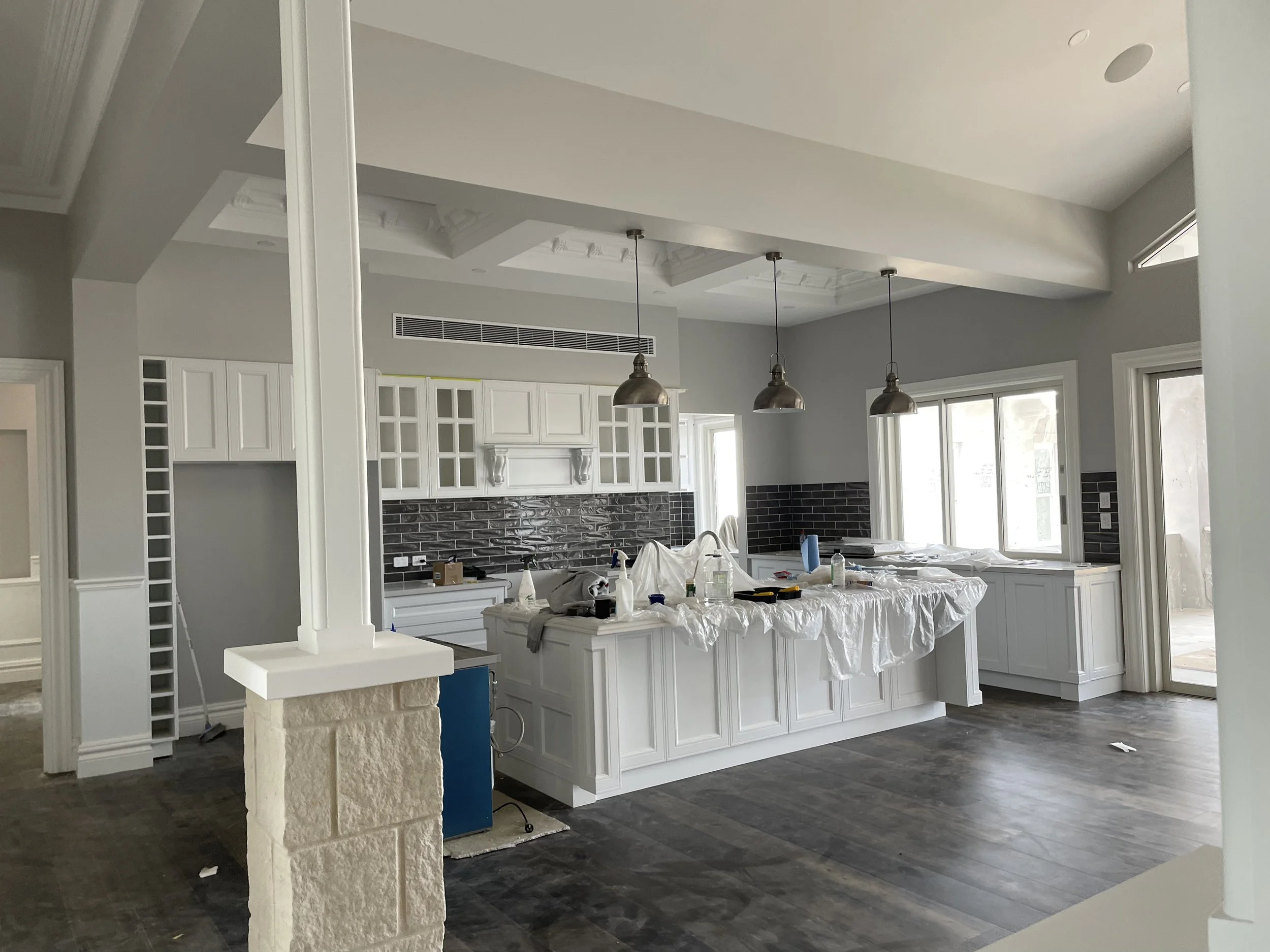 Modern kitchen under construction with white cabinetry, black tile backsplash, covered countertops, and pendant lighting.