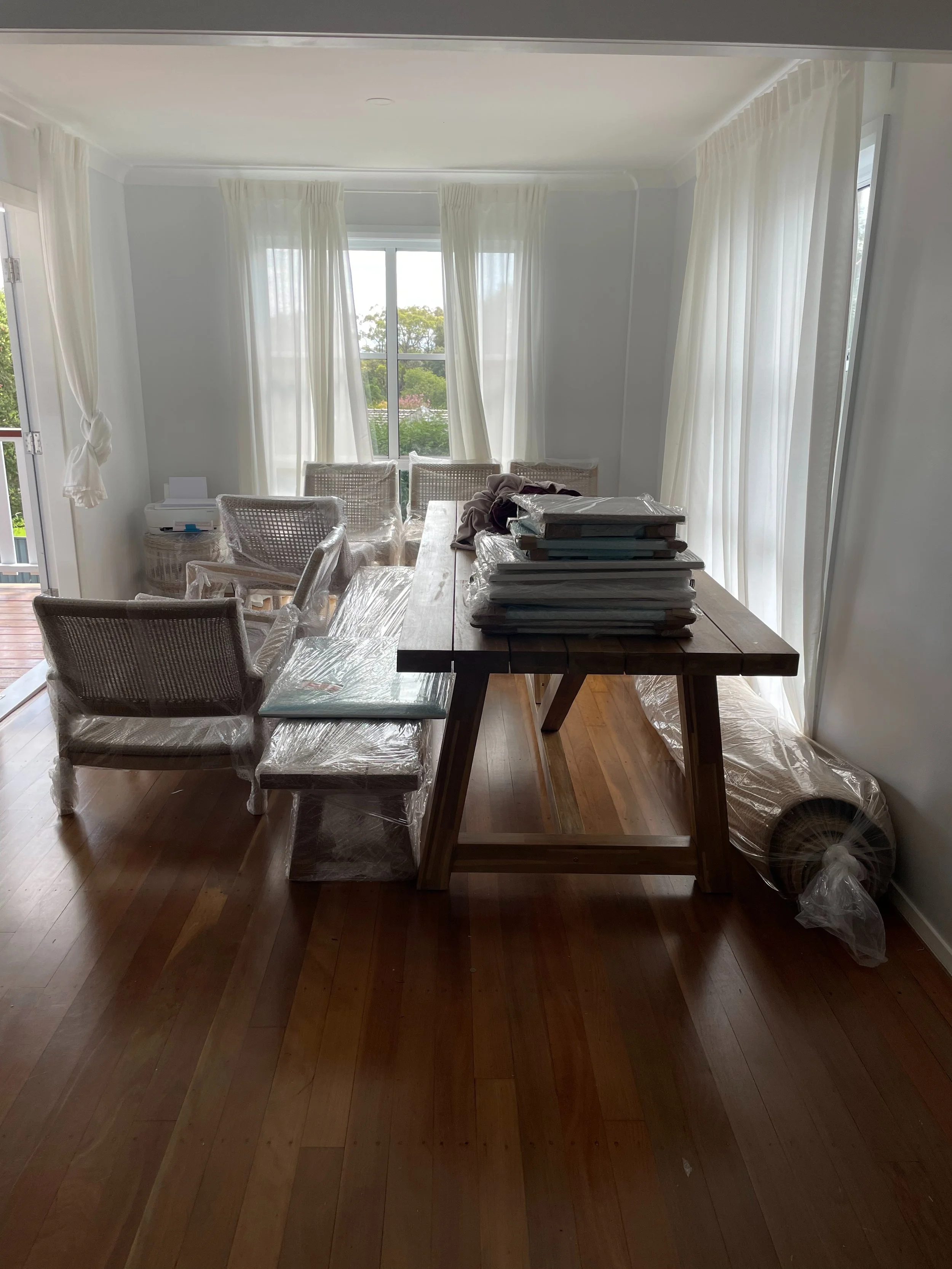 Dining room with wooden floor, table, and chairs covered in plastic, white curtains, and large windows.