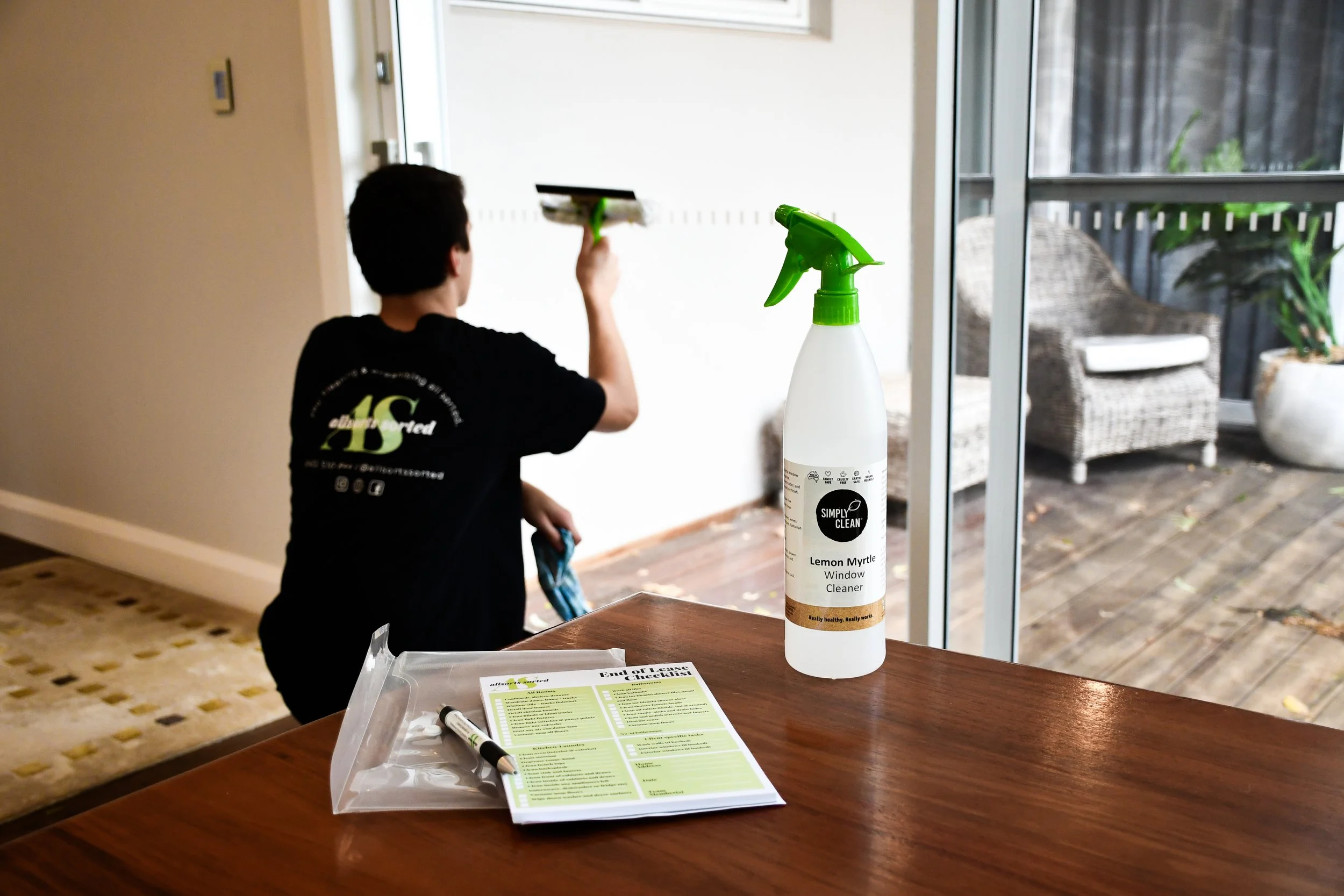 A person cleaning a glass door from inside a house, with a bottle of Lemon Myrtle window cleaner, a marker, and a cleaning checklist on a table in the foreground.