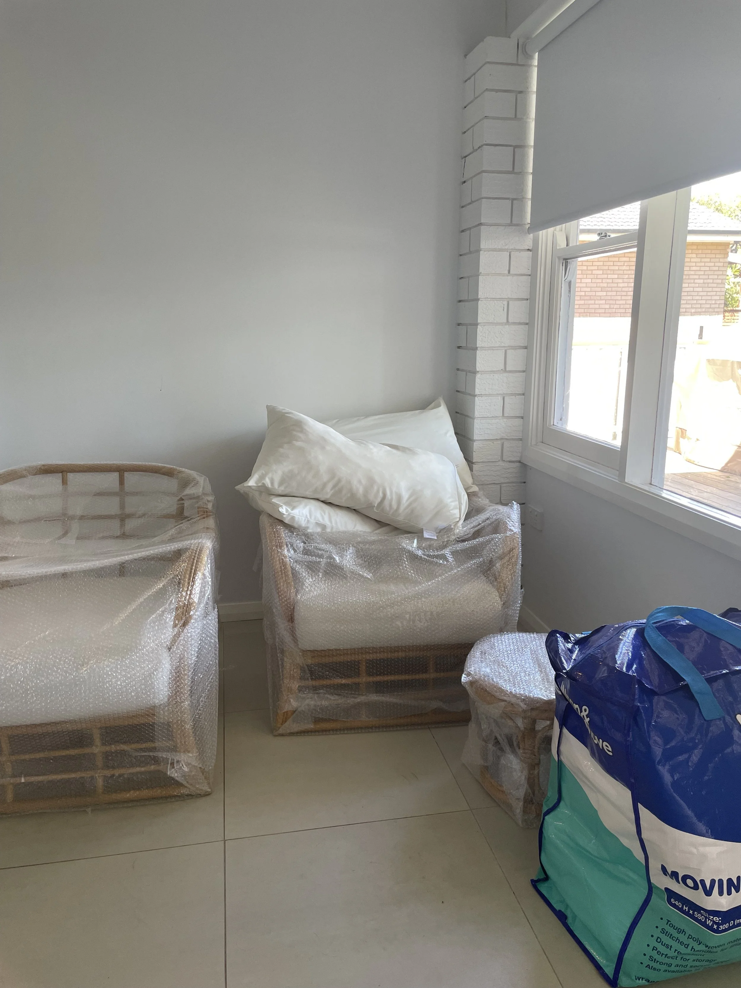 Furniture wrapped in bubble wrap with a large blue and white moving bag in a corner room with white walls and a window.