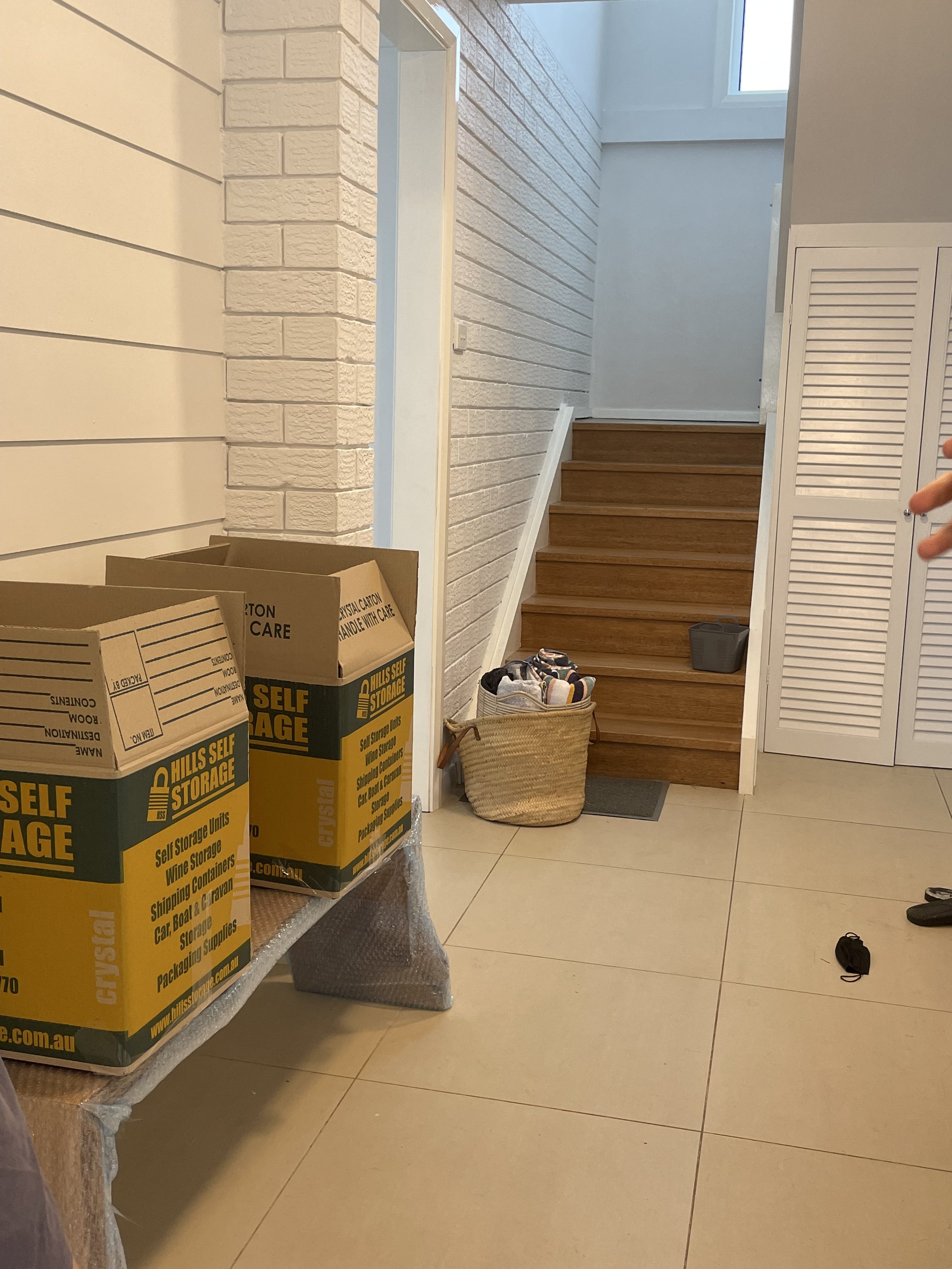 Stacks of cardboard boxes labeled "self storage" on a plastic-covered table in a hallway with white brick walls. Wooden stairs lead upward, and a woven basket with items is on the floor.