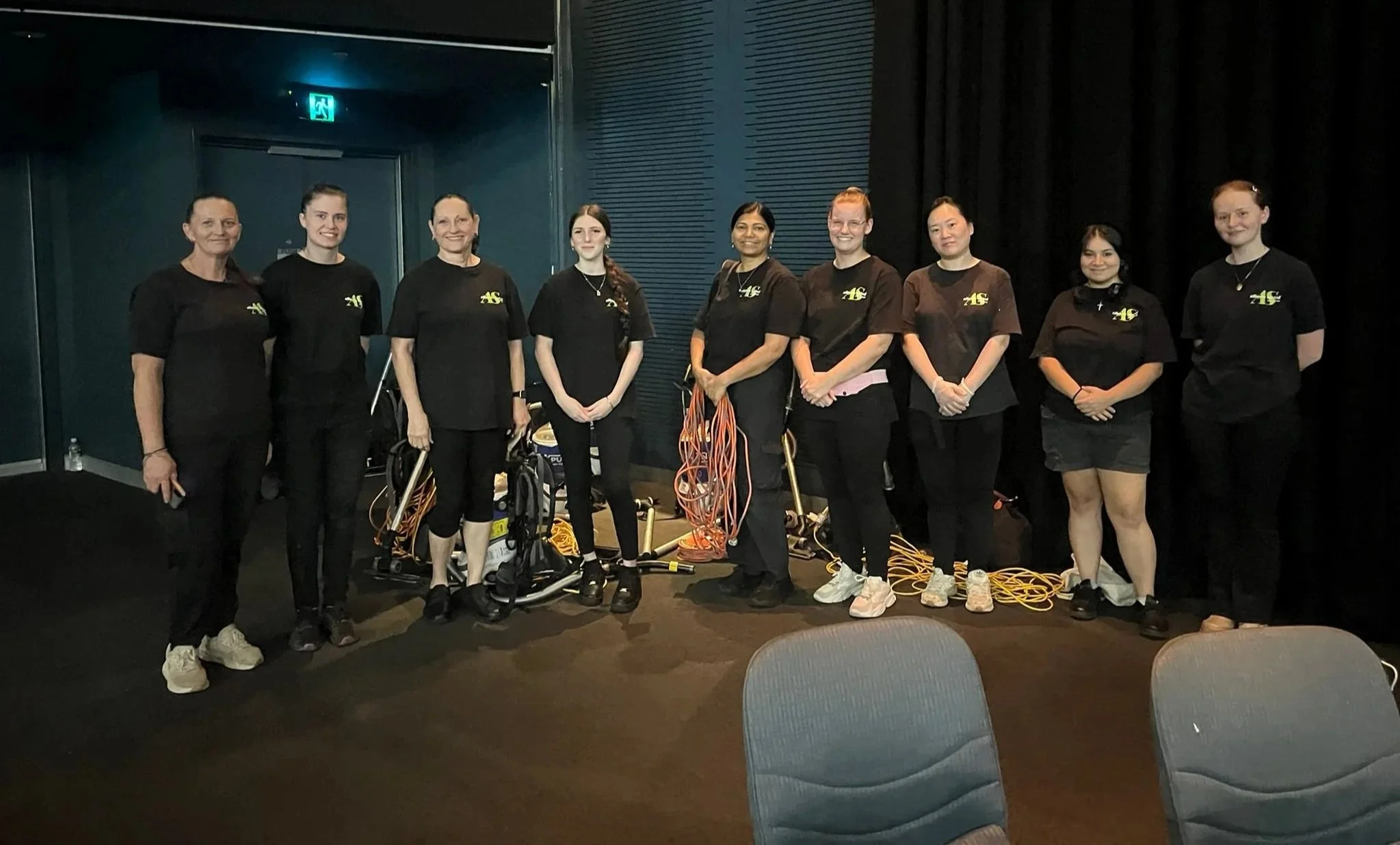 Group of nine women standing together in a room, wearing black t-shirts with yellow logos, some with cables and equipment around them.