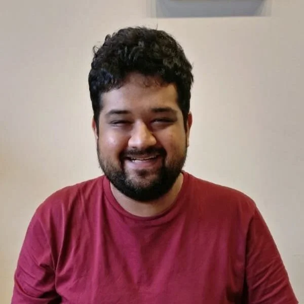 Man with curly hair and beard wearing a red shirt smiling while sitting indoors.